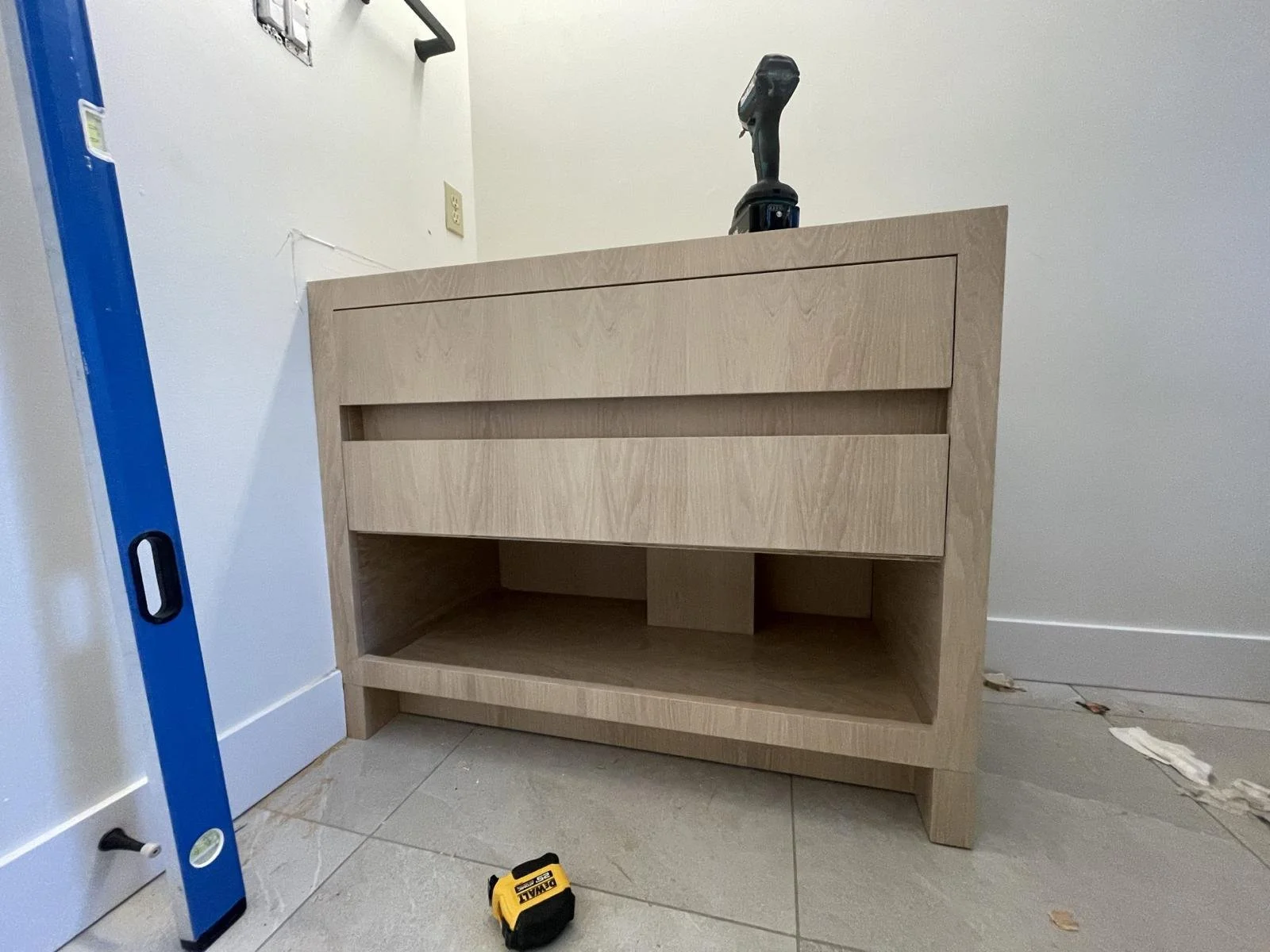 Unfinished wooden cabinet with two drawers and an open shelf at the bottom, in a room with construction tools nearby.