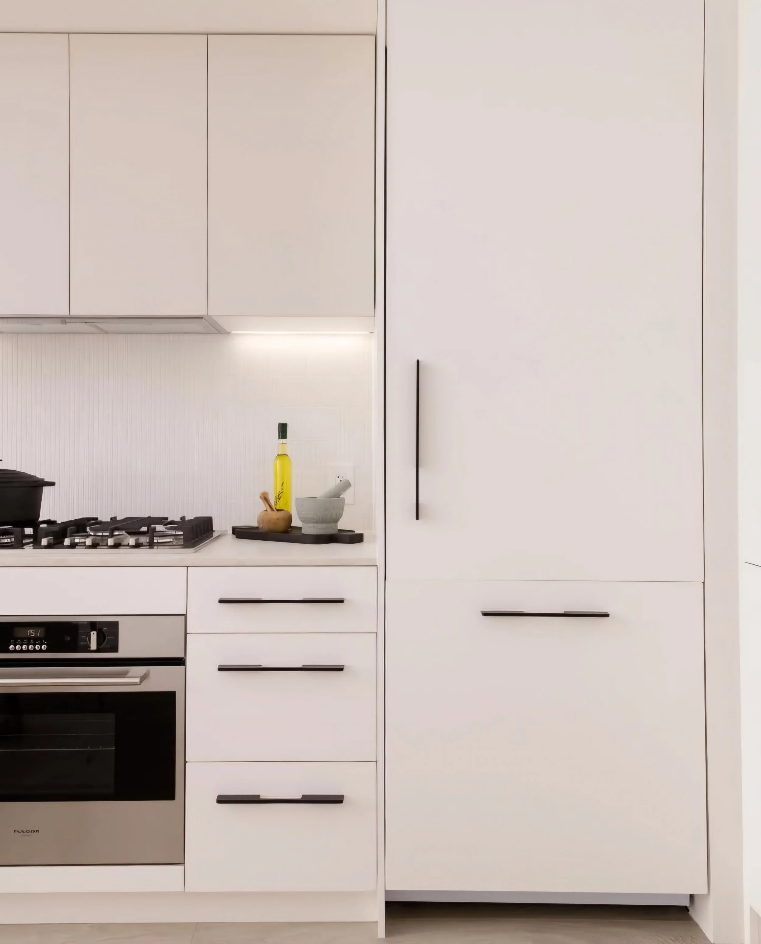 White kitchen cabinets and drawers with black handles, a built-in oven, a stovetop, a small bowl, a green-and-yellow bottle, and a mortar and pestle on the countertop.
