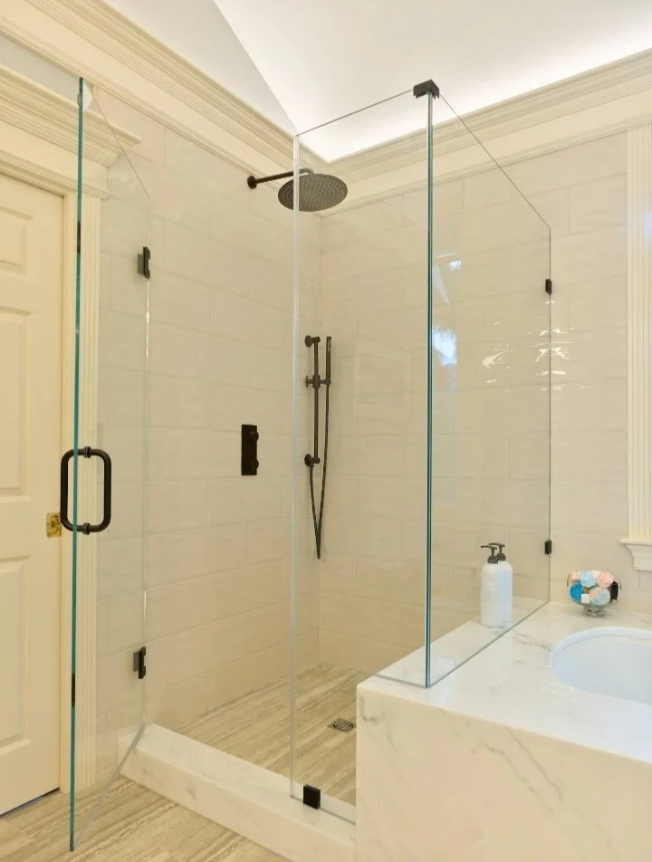Modern shower area with clear glass doors, a rainfall showerhead, and a hand-held shower fixture inside a bathroom with beige tiled walls and marble countertop.