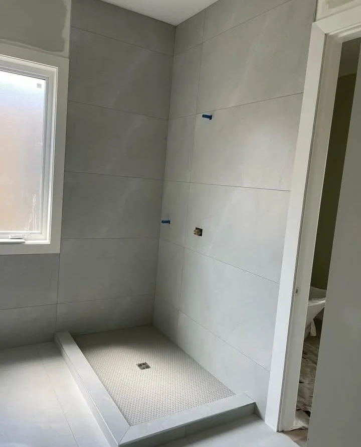 Empty bathroom shower area with tiled walls, a window, and plumbing fixtures installed. The shower base is positioned on the floor, awaiting finishing touches.