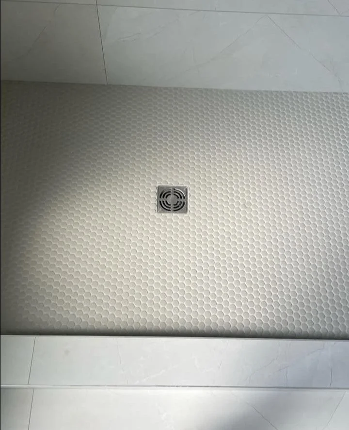 Close-up of a tiled shower floor with a square drain in the center, surrounded by small hexagonal tiles.