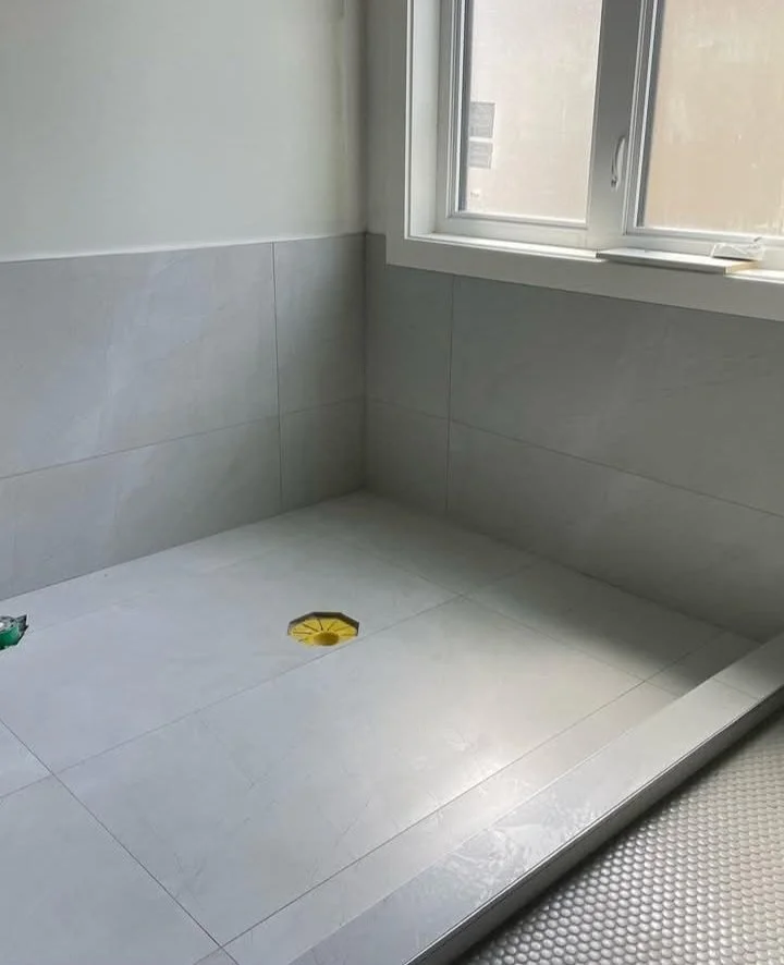 Empty bathroom shower area with gray tiles, a drain on the floor, and a window with a white frame.