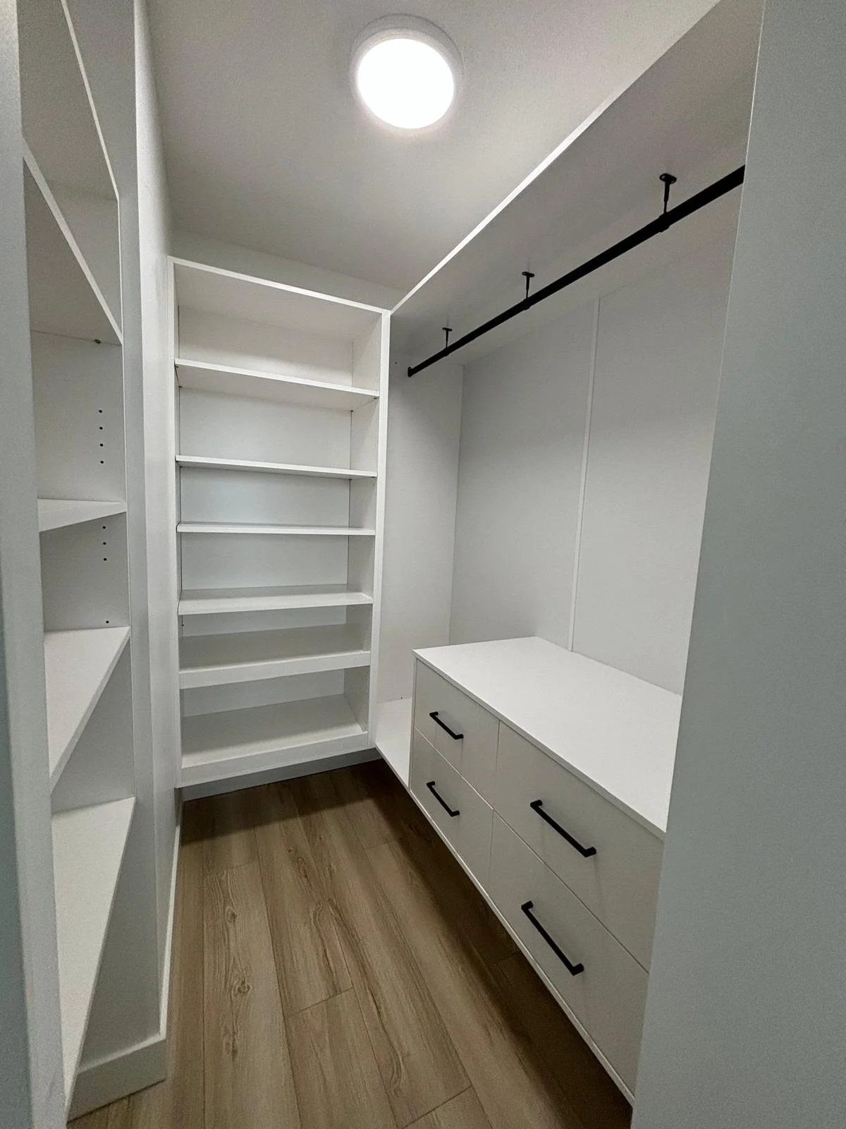 Empty walk-in closet with white shelving and drawers, a hanging rod, and wood flooring.