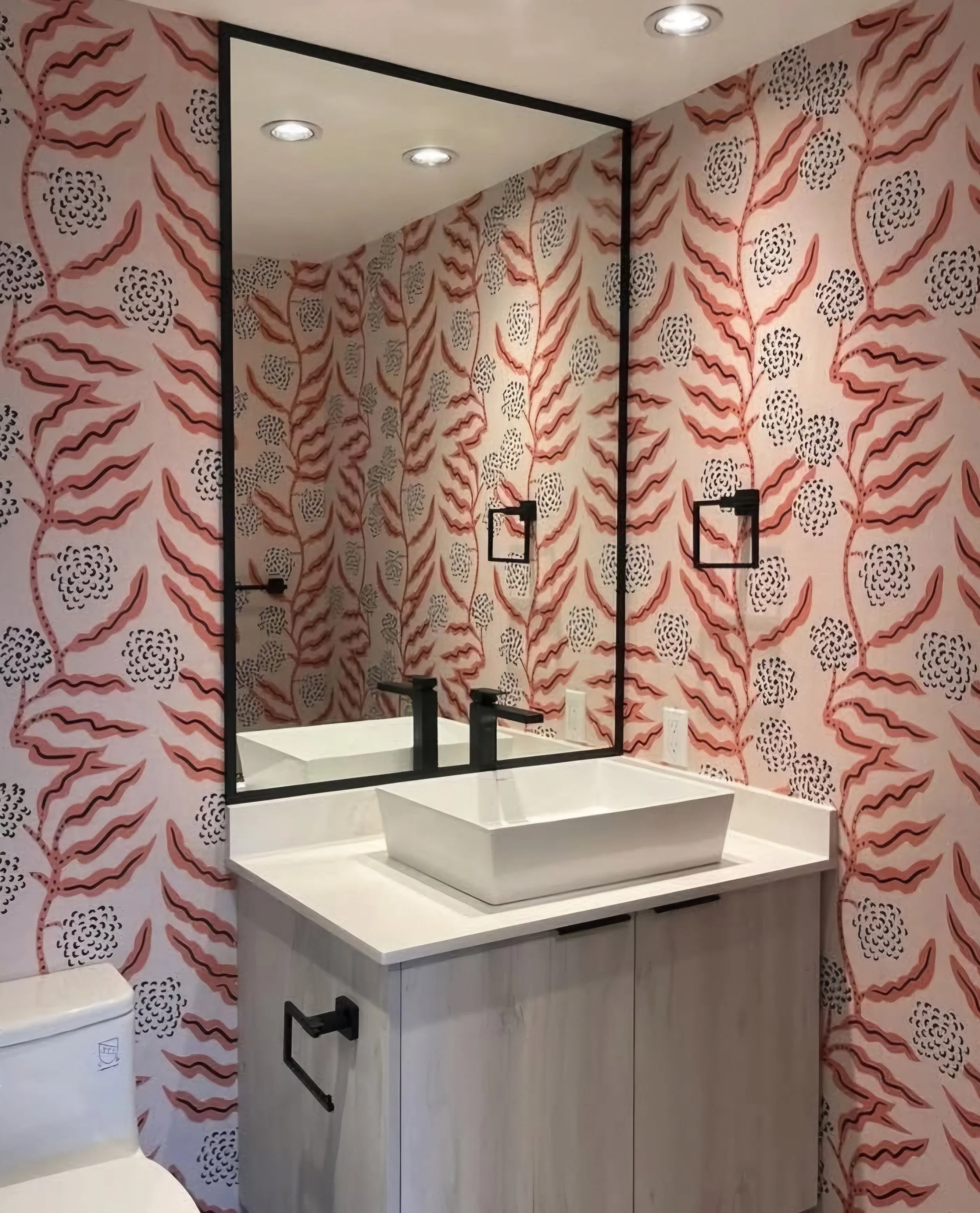 A bathroom with pink and black floral patterned wallpaper, a large mirror, a white rectangular sink, black fixtures, and a light-colored cabinet underneath the sink.