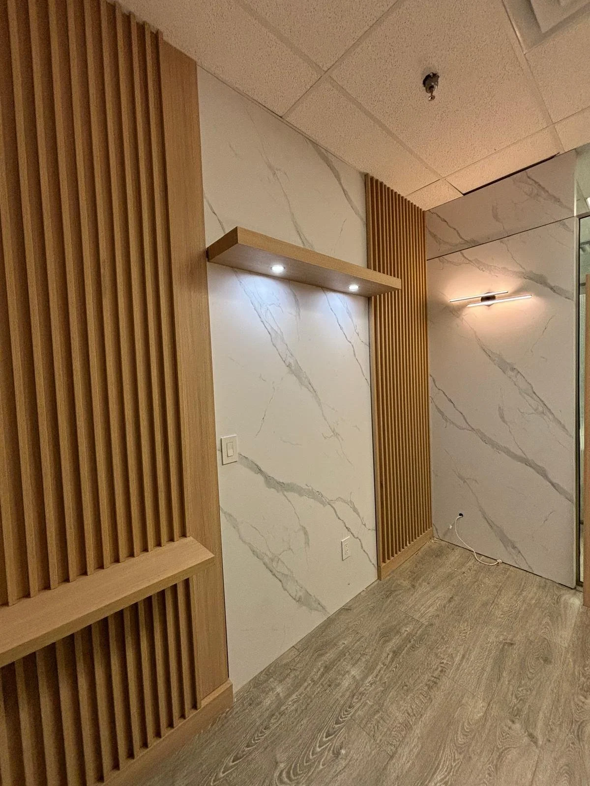 Interior corner of a room with white marble walls with gray veins, wooden vertical slats on two sides, a floating wooden shelf with built-in lights, and a wall-mounted light fixture, with a hardwood floor and an electrical outlet on the wall.