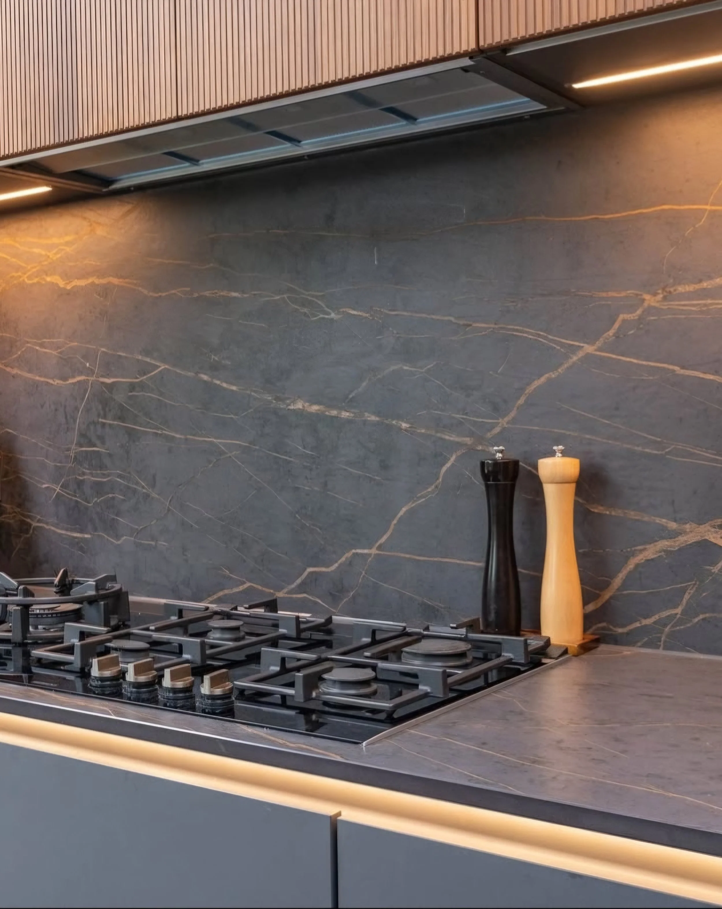 Modern kitchen stove with a black and a light wood salt or pepper mill on a dark marble countertop against a matching dark marble backsplash.