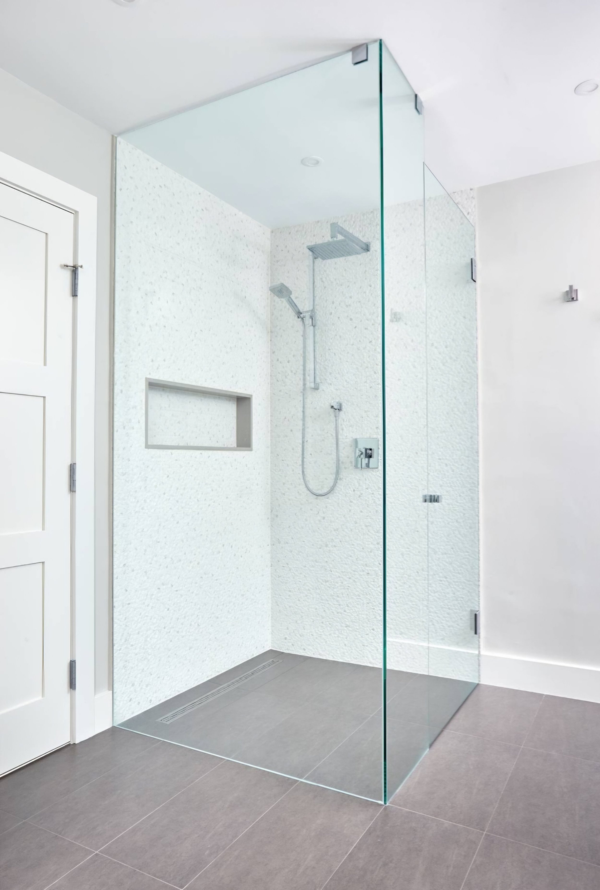Modern walk-in shower with clear glass walls, rainfall showerhead, and textured white wall tiles, adjacent to a white door on a gray-tiled floor.