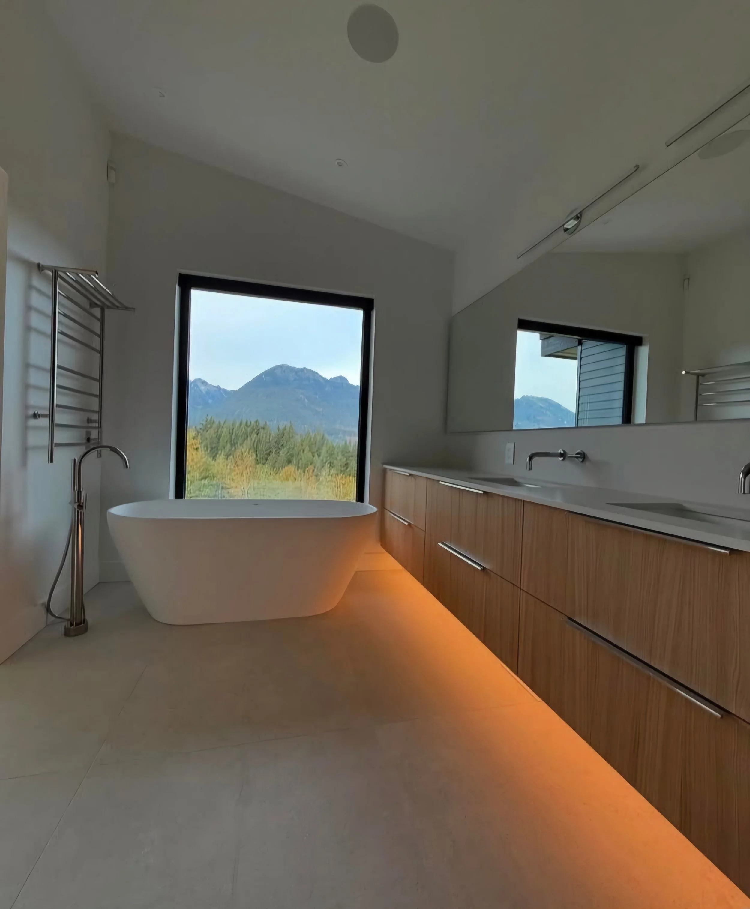Modern bathroom with a freestanding bathtub, wooden vanity with double sinks, large window showing mountains, and neutral-colored flooring.