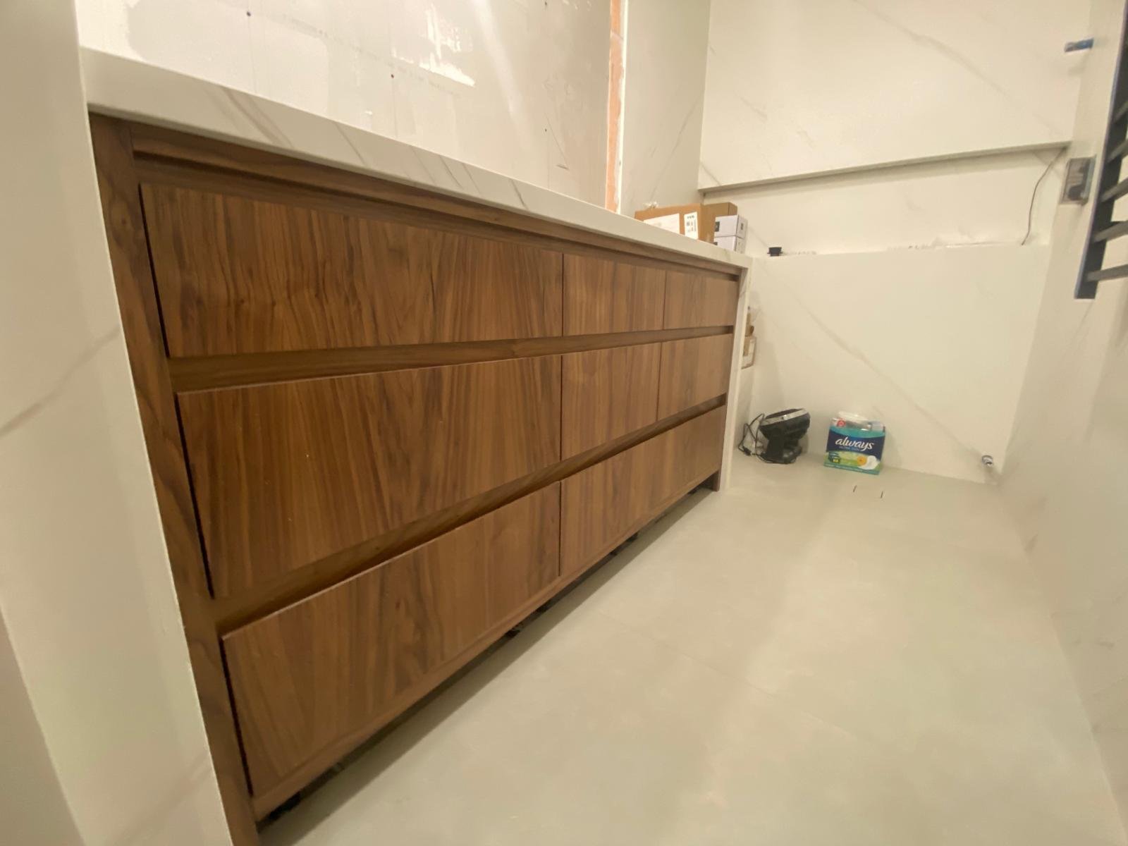 Wooden dresser in a room with beige tiled floor and white walls. Items including a box of Always pads and a black bag are on the floor.
