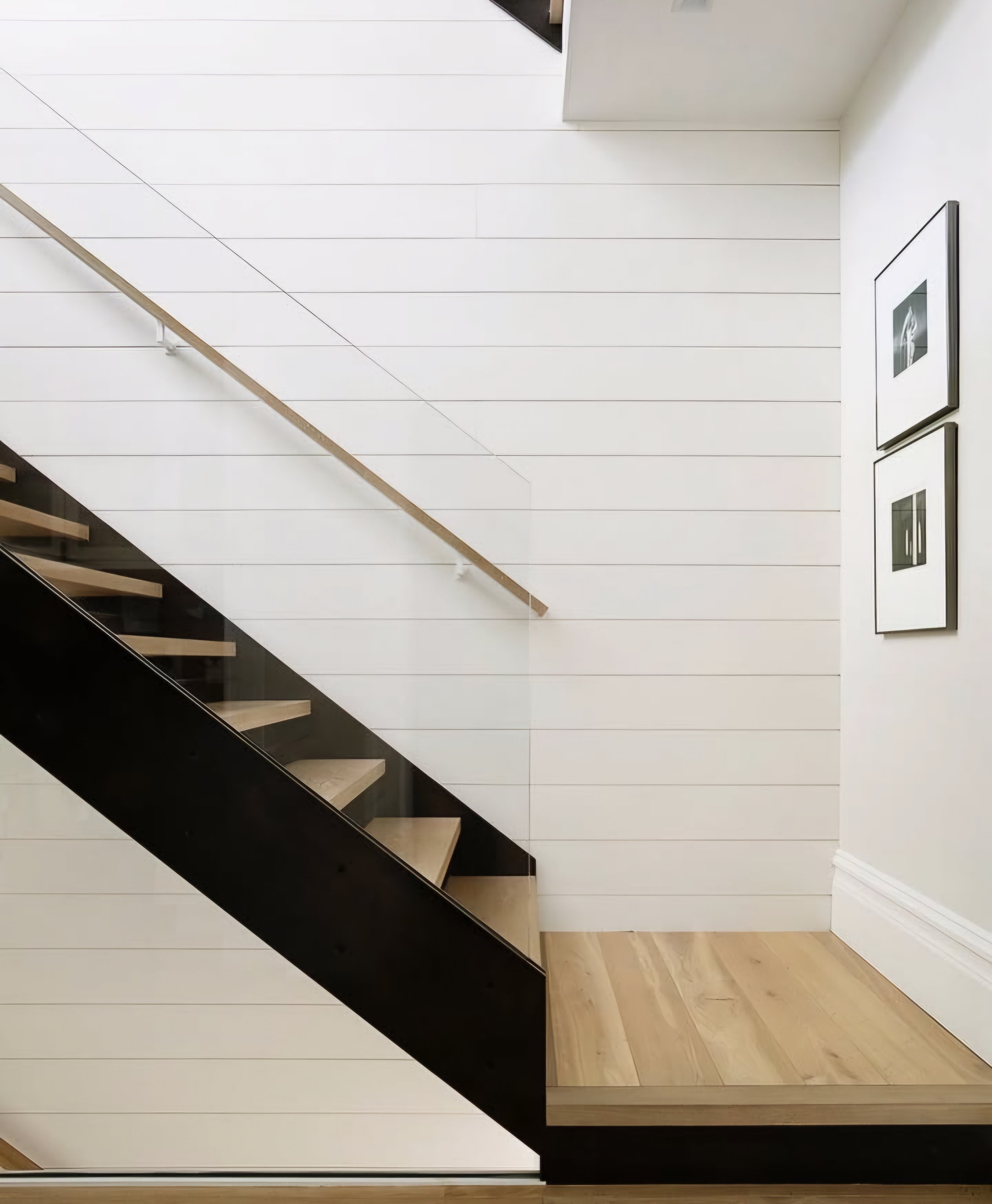 Interior view of a modern staircase with light-colored wooden steps, a glass railing, and white horizontal panel walls decorated with black and white framed artwork.