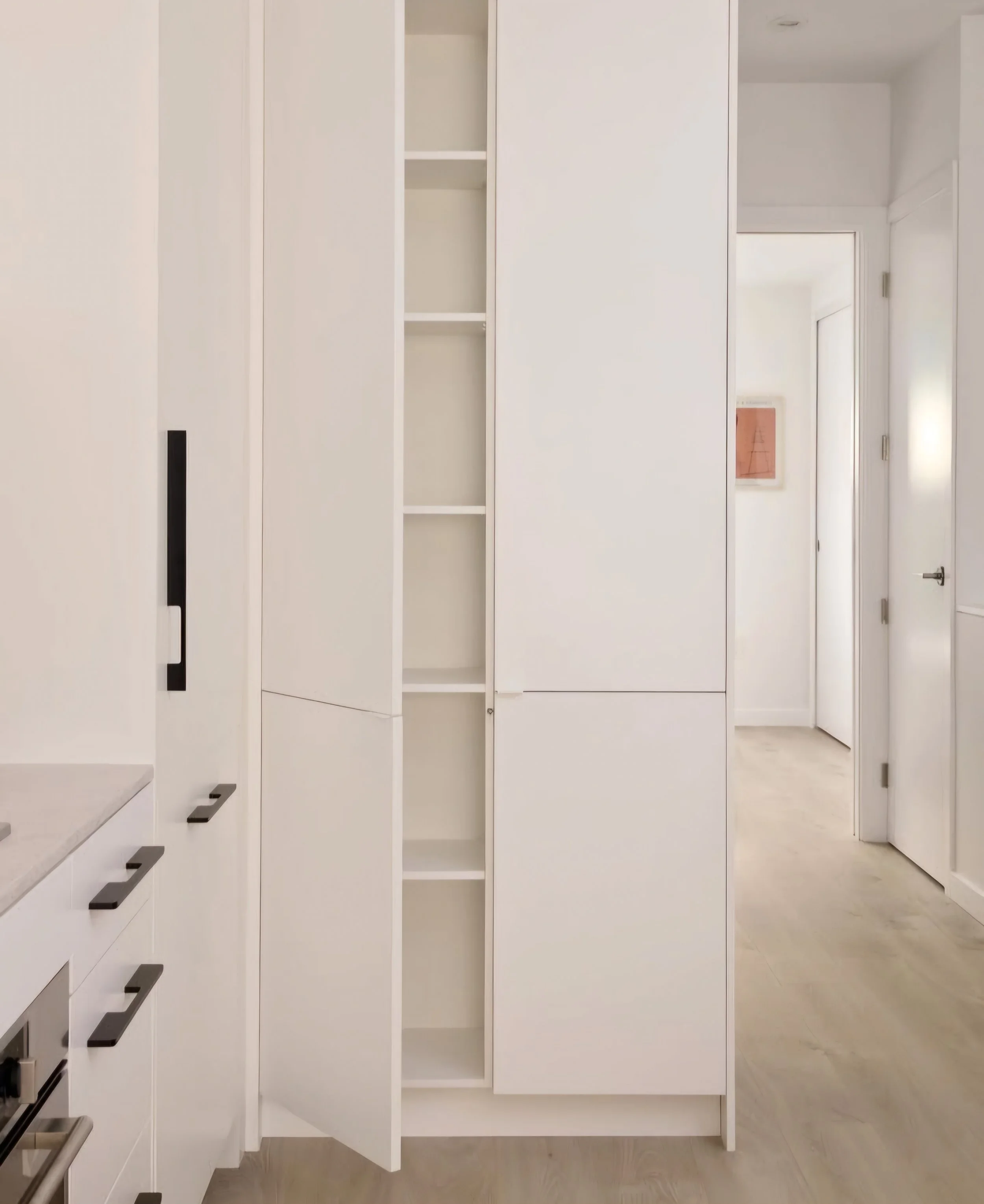 Open white kitchen cabinet with shelves, adjacent to other closed cabinets in a modern kitchen with light wood flooring.