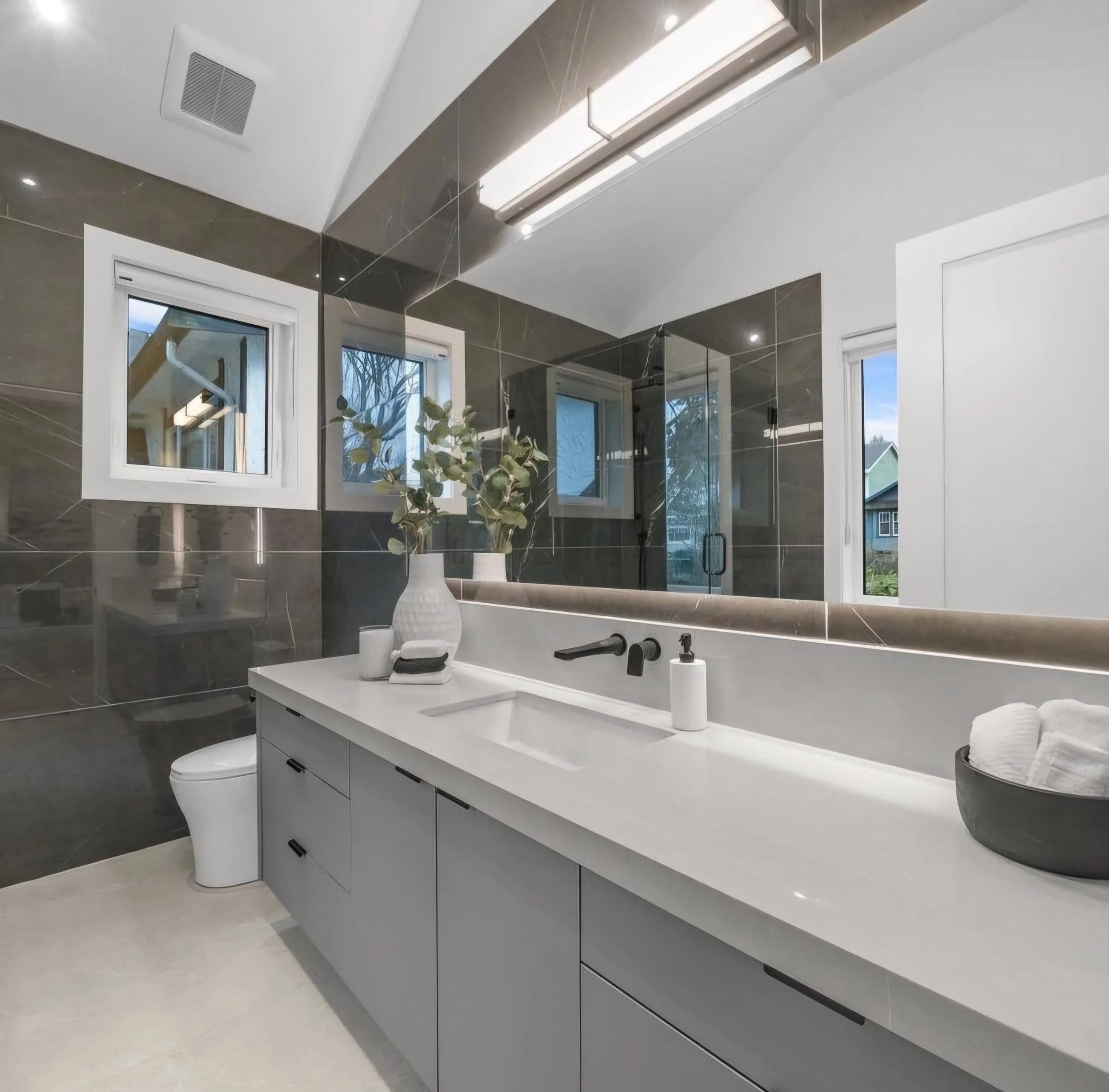 Modern bathroom with white vanity, black fixtures, large mirror, dark tiled walls, and small windows.