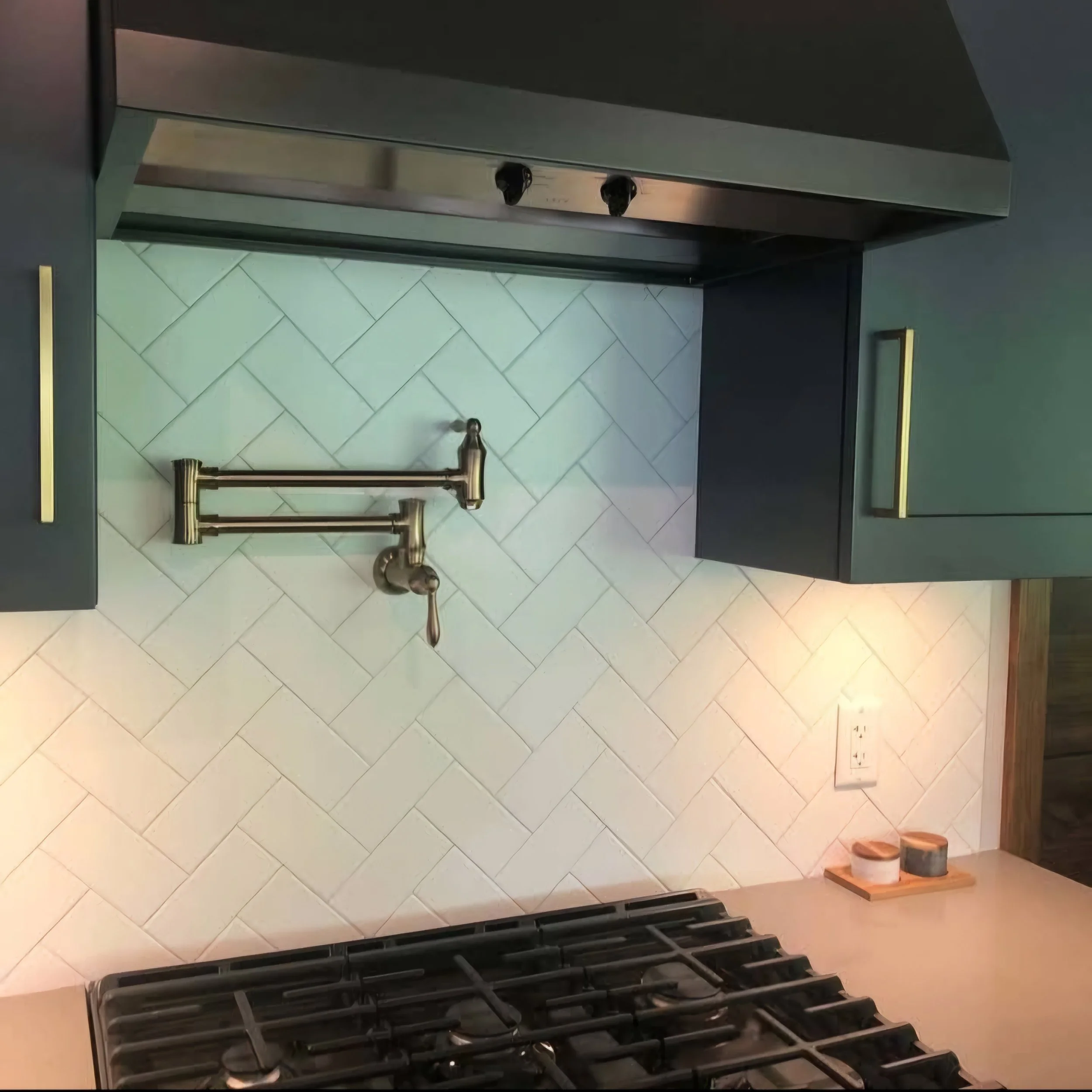 Kitchen wall with a white herringbone tile backsplash, dark gray upper cabinets with gold handles, a stainless steel range hood, a gas stove, a pot filler faucet, and small containers on a beige countertop.