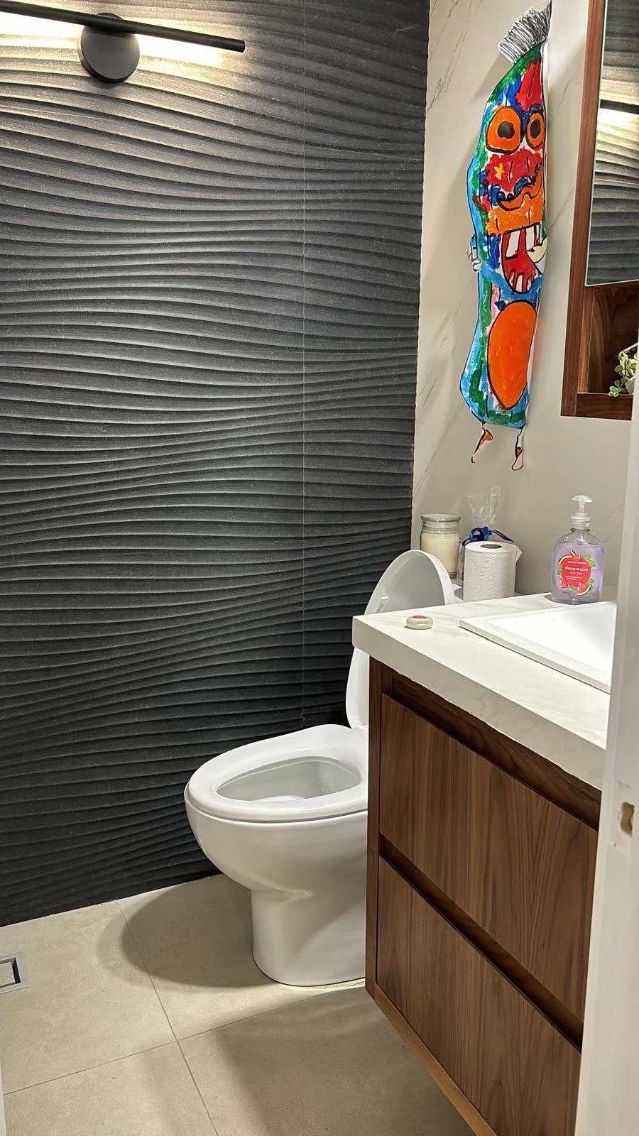 A small bathroom with a black textured wall, a white toilet, a white sink with a wooden cabinet underneath, and a colorful decorative fish wall hanging.