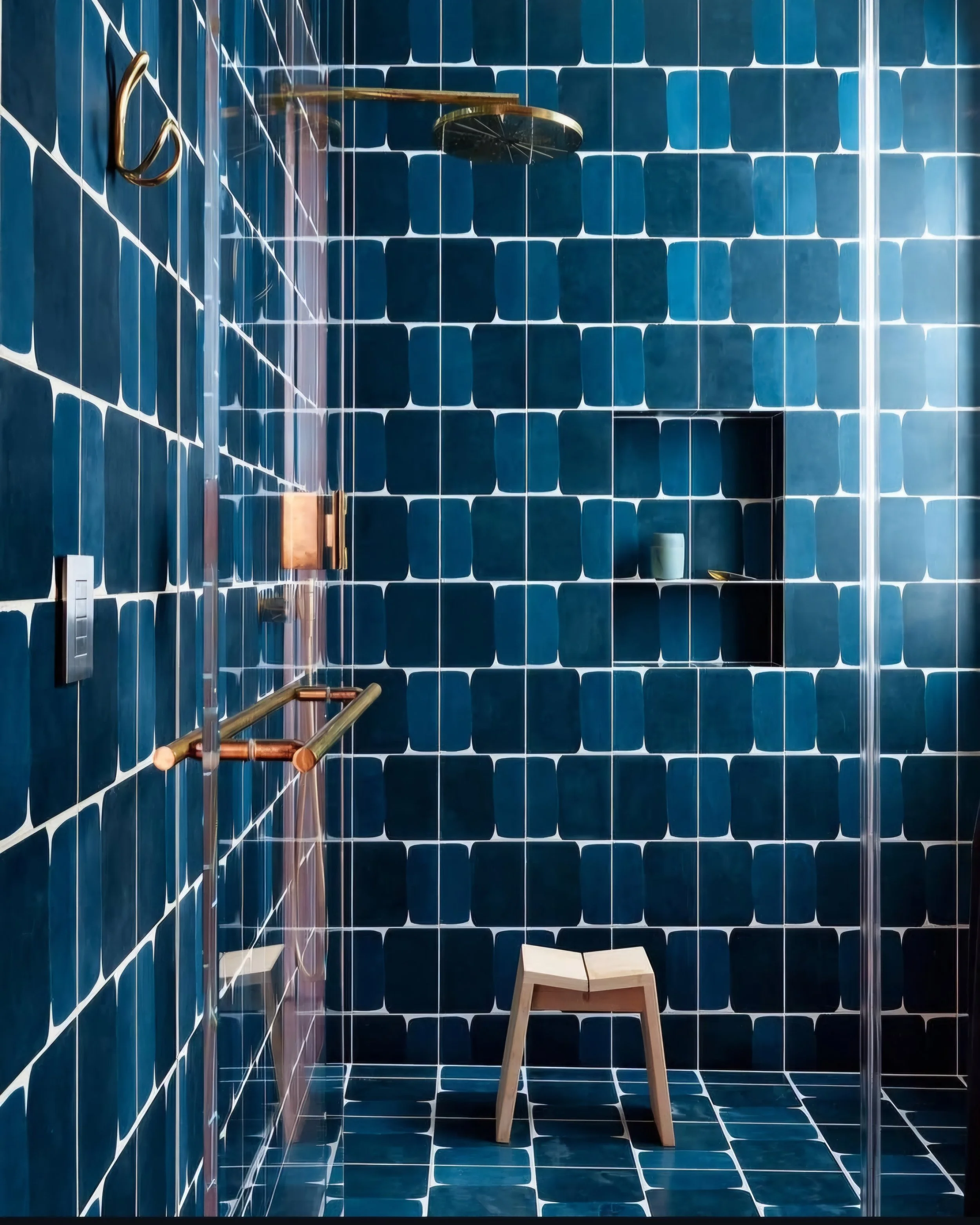 Modern shower area with dark blue square tiles, a round ceiling shower, a small built-in shelf with a cup, a brass grab bar, a door handle, a support rail, and a wooden stool.