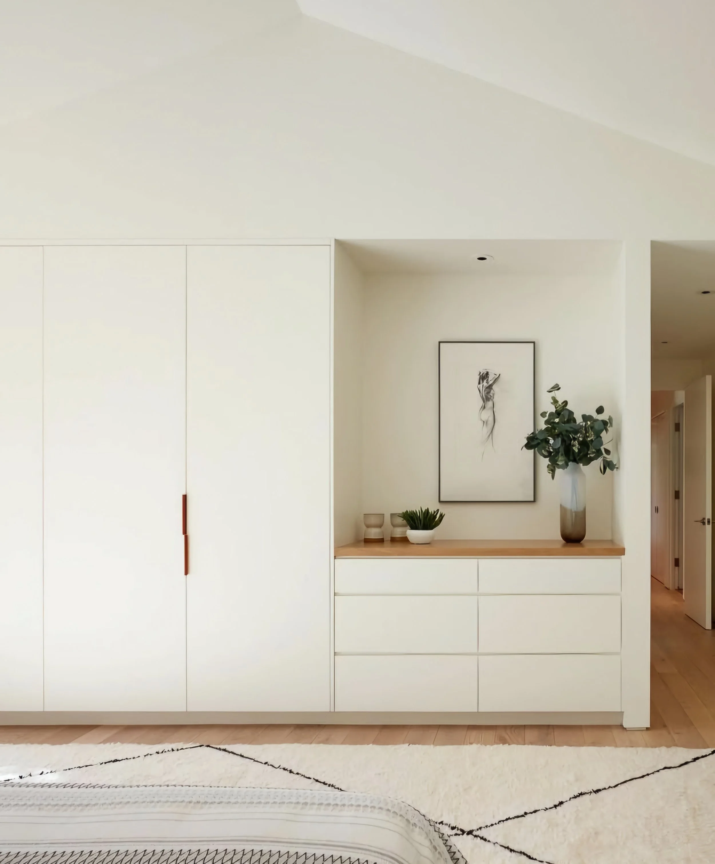 Minimalist bedroom dresser area with framed art, a tall vase with green foliage, small bowls, and a potted plant on a wooden surface.