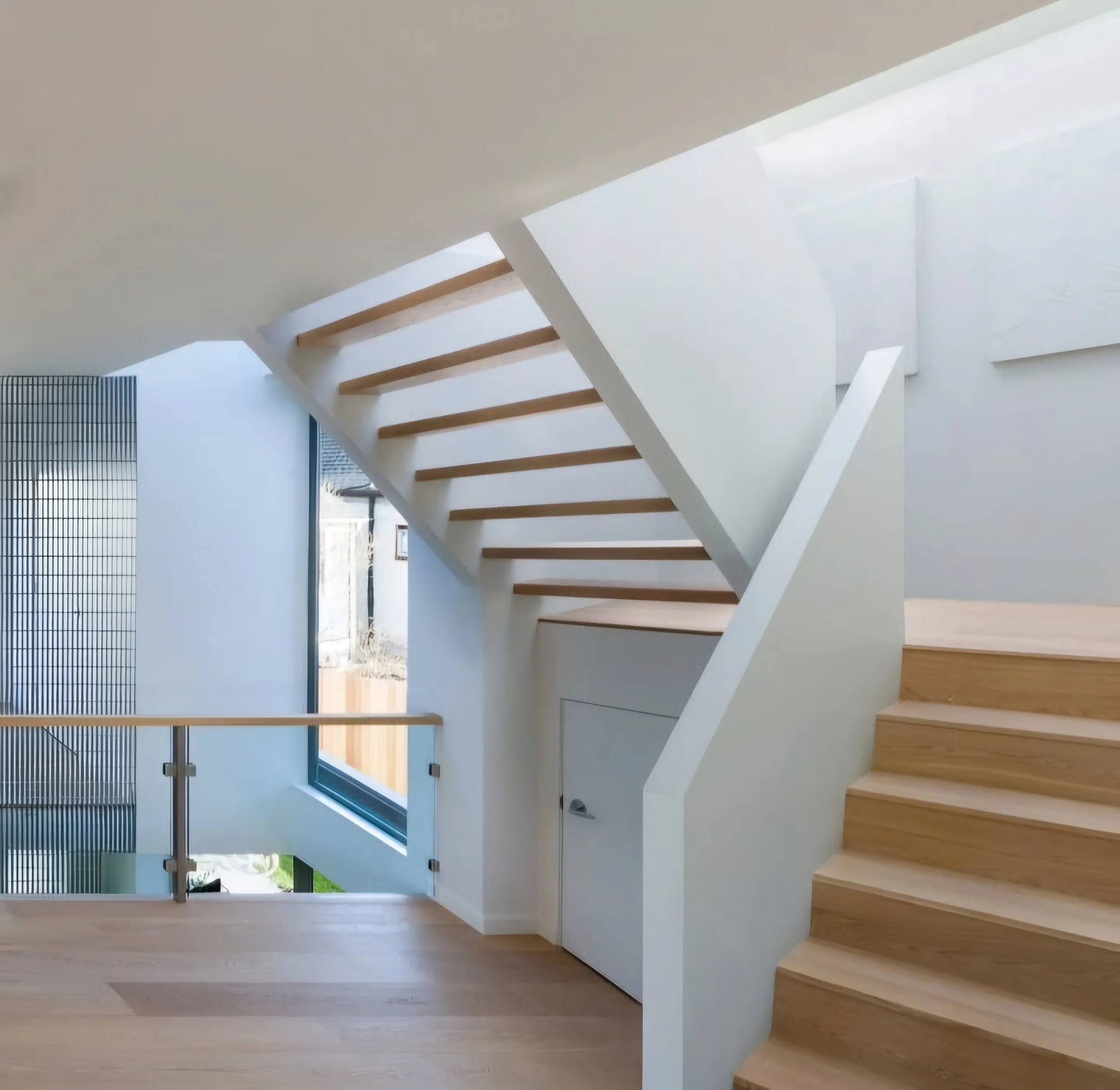 Interior view of a modern staircase with wooden steps and a split-level design, featuring glass and wood railing, in a home with large windows and natural light.
