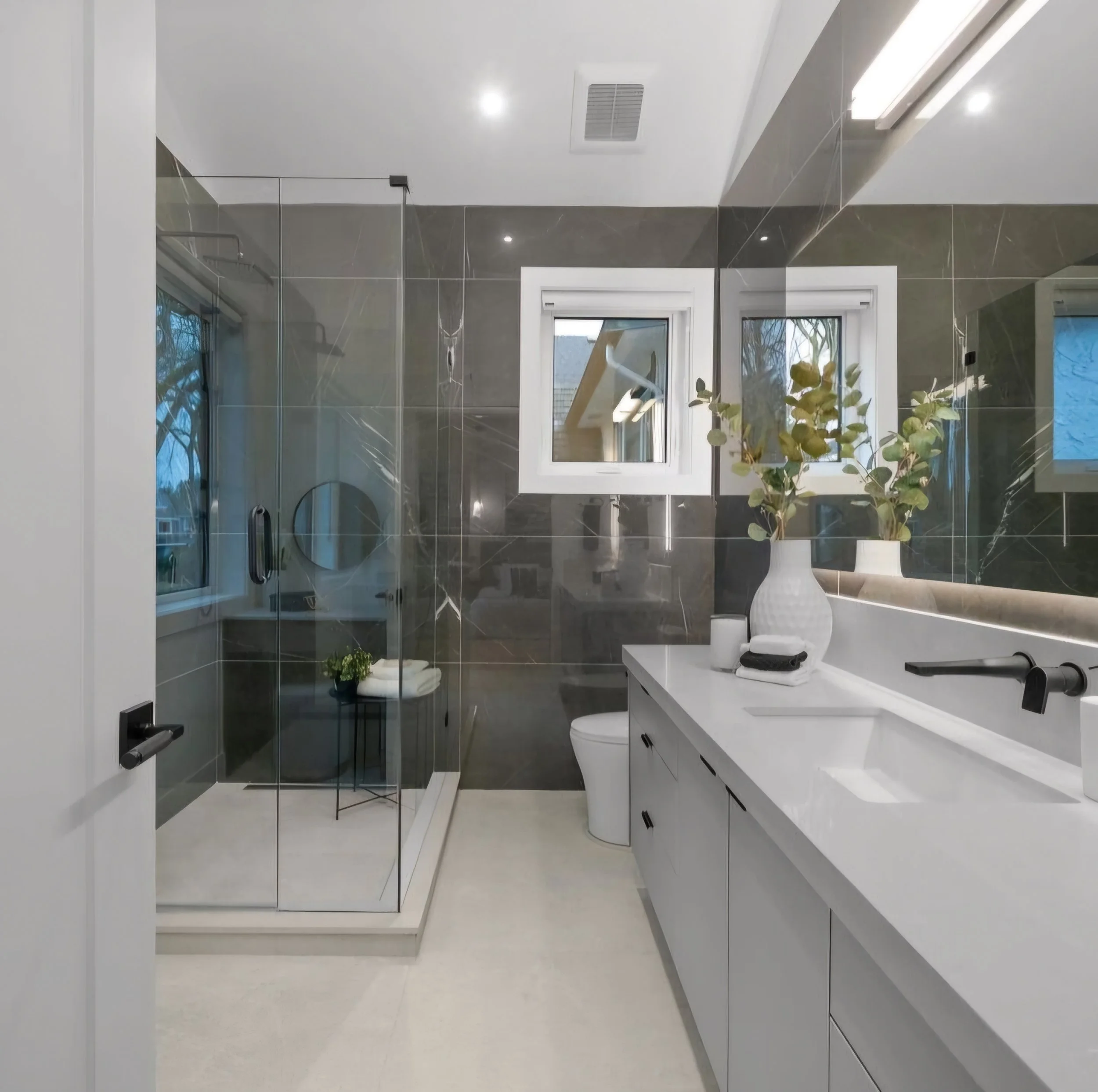 Modern bathroom with gray marble walls, a white vanity with black handles, a large mirror, and two small windows. There is a glass shower enclosure with a small black side table holding towels and a plant.
