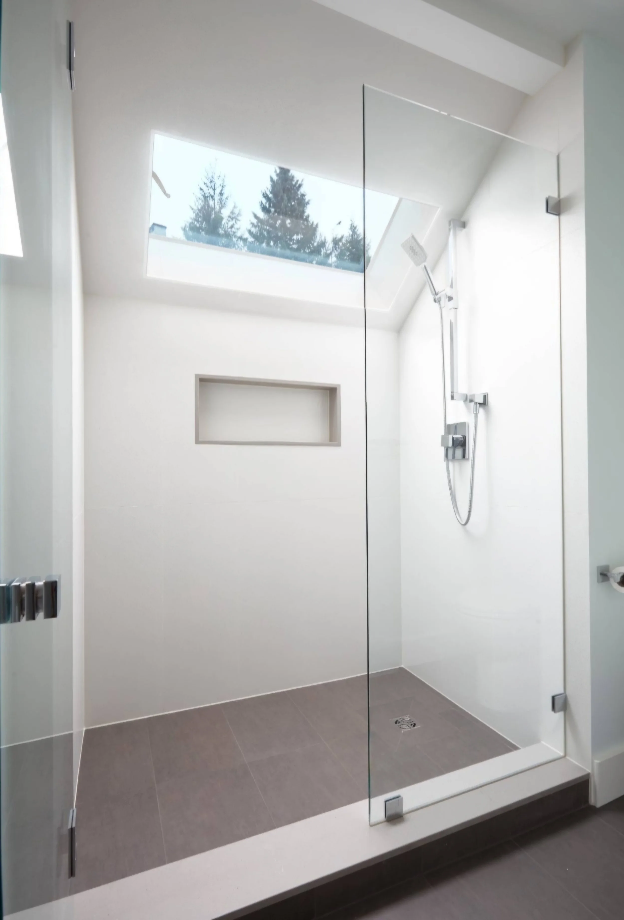 Modern walk-in shower with a large rectangular skylight, white walls, a built-in shelf, and gray floor tiles.