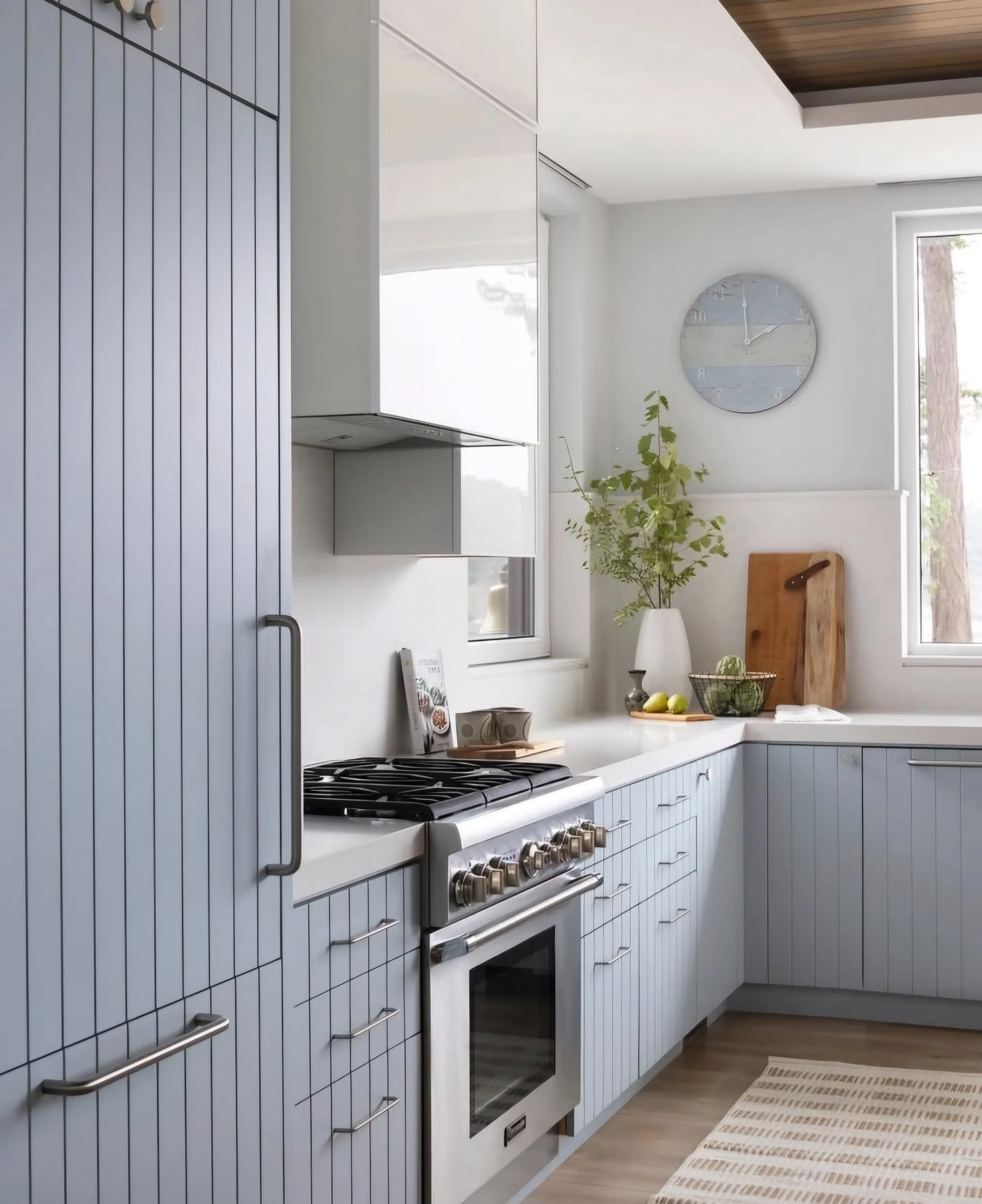 Modern kitchen with light gray vertical-paneled cabinets, a stainless steel oven and stove, white countertops, a potted plant, a wooden cutting board, a bowl of green vegetables, lemons, and a clock on the wall.