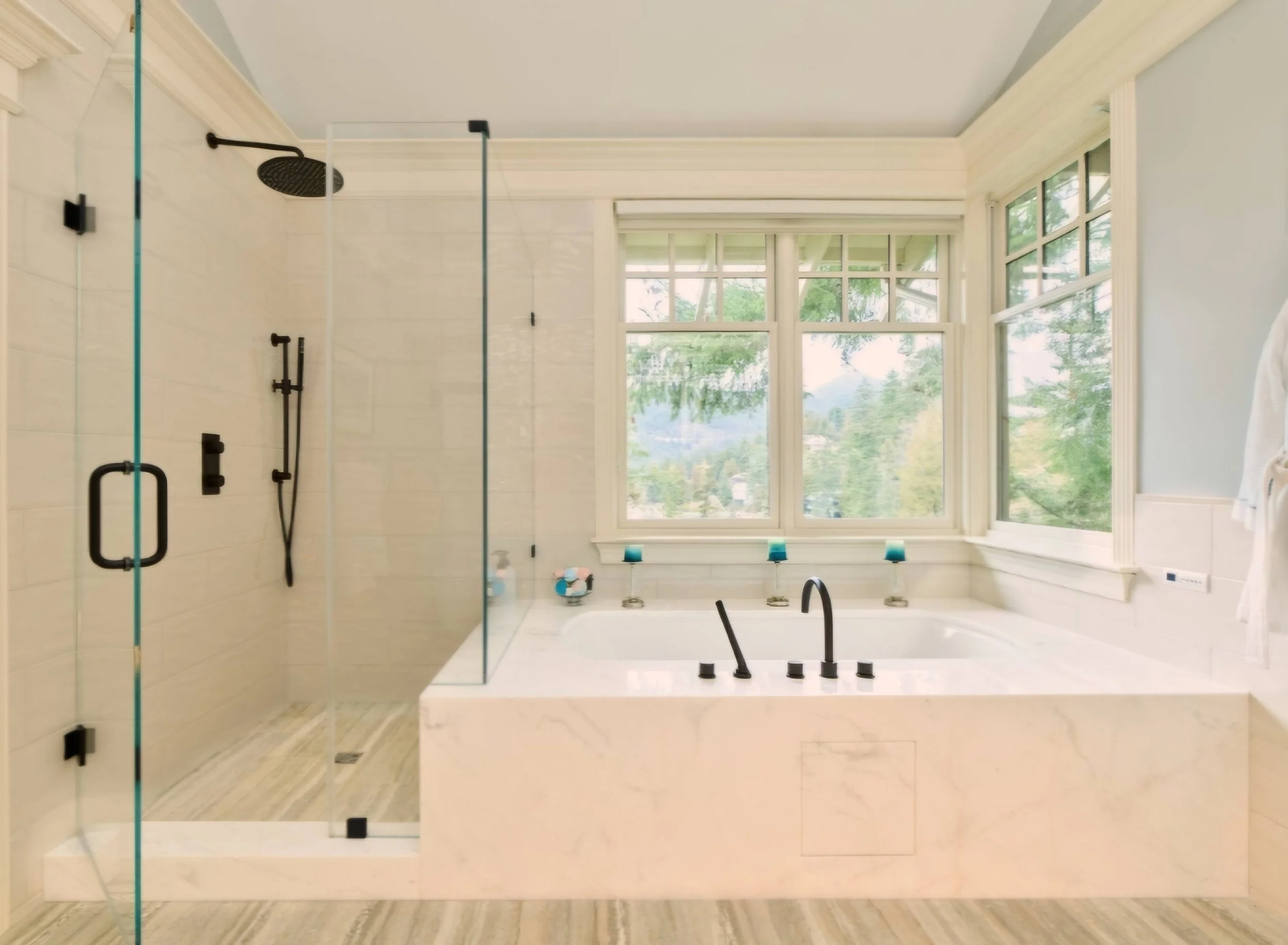 A modern bathroom with a large walk-in shower with glass doors and black fixtures, next to a bathtub with black fixtures, surrounded by large windows showing a view of trees and mountains.