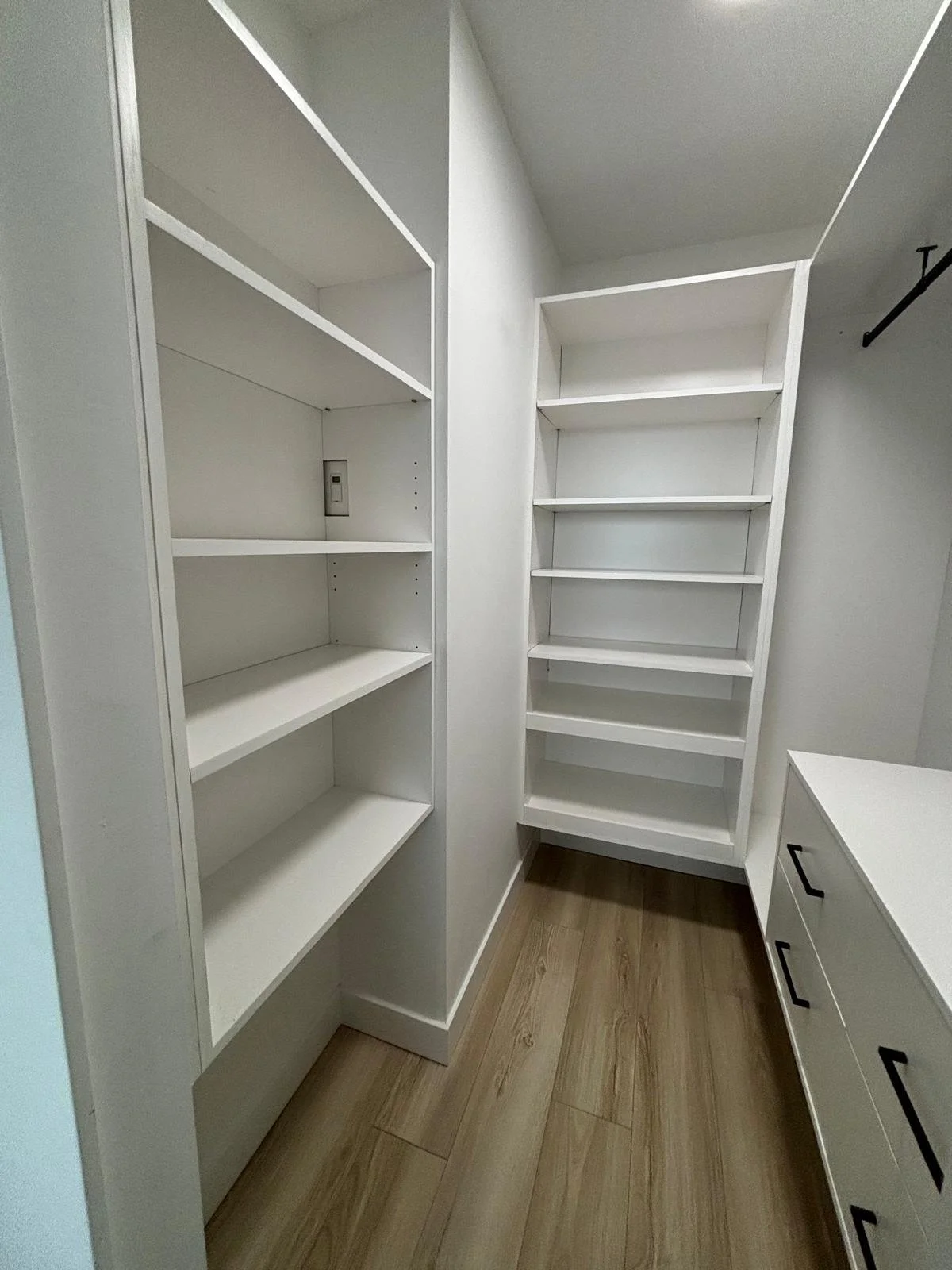 Empty walk-in closet with white shelves and drawers, wooden floor, and a wall-mounted hook.