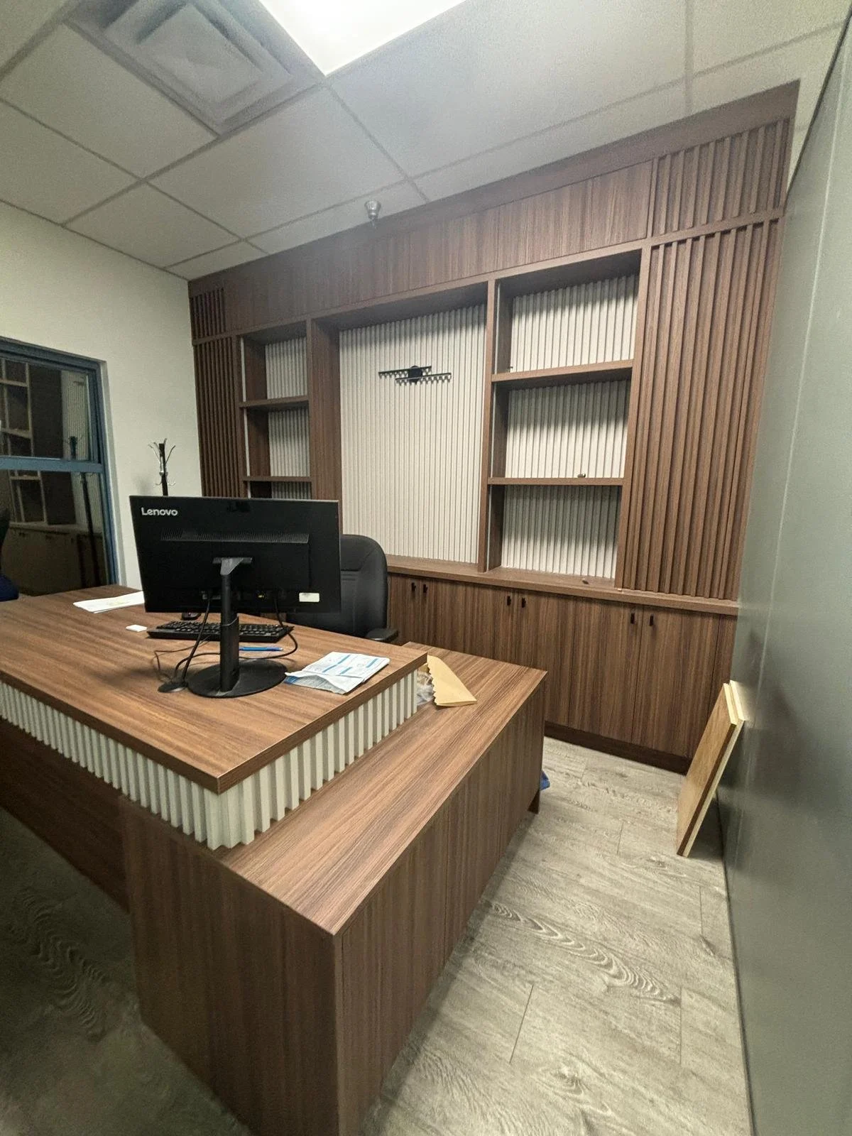 Office with a wooden desk, a computer monitor, paperwork, and wood shelving on the wall.