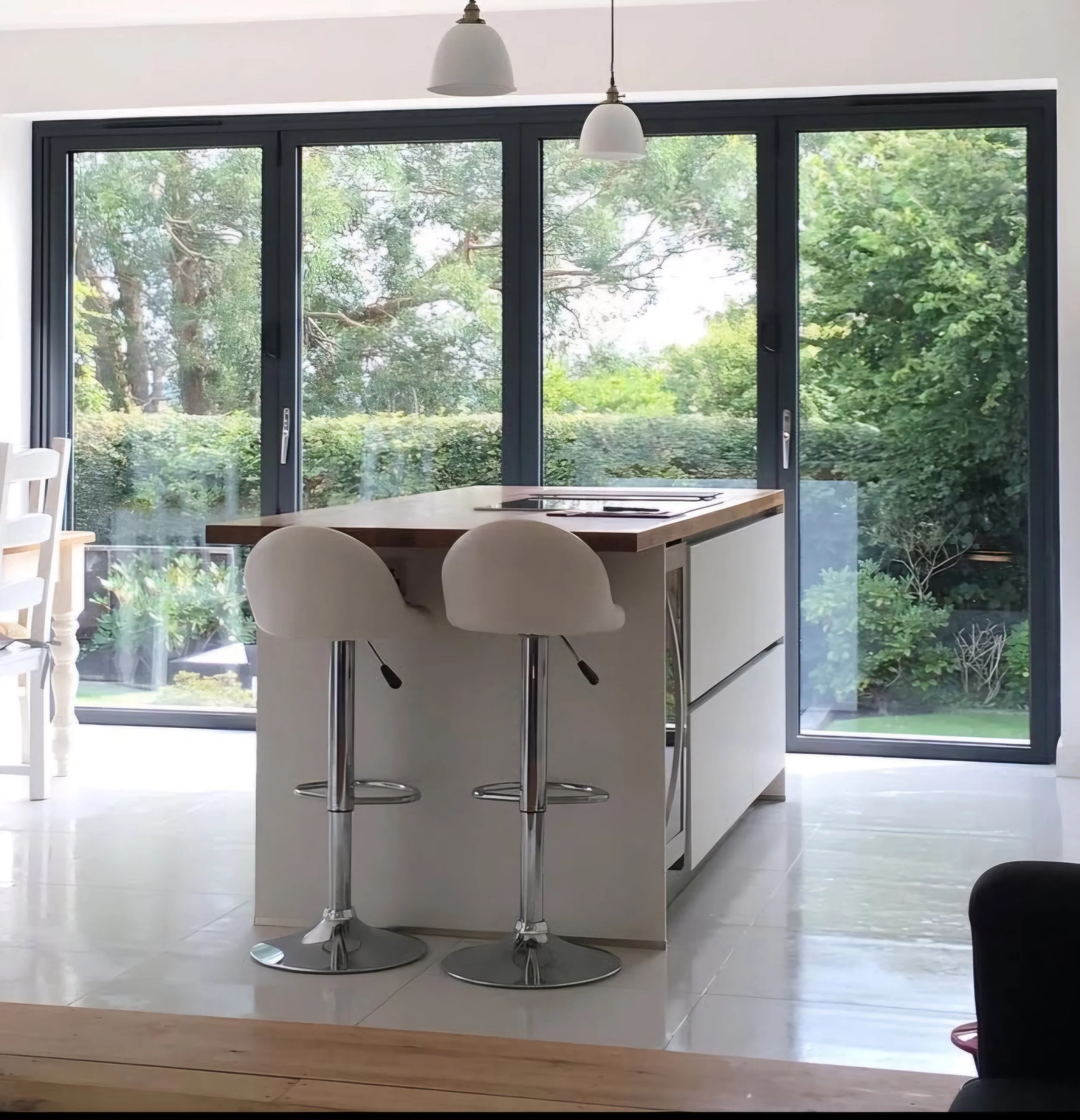 Modern kitchen with white island, two white barstools, large sliding glass doors showing green trees outside, pendant lights hanging from the ceiling.