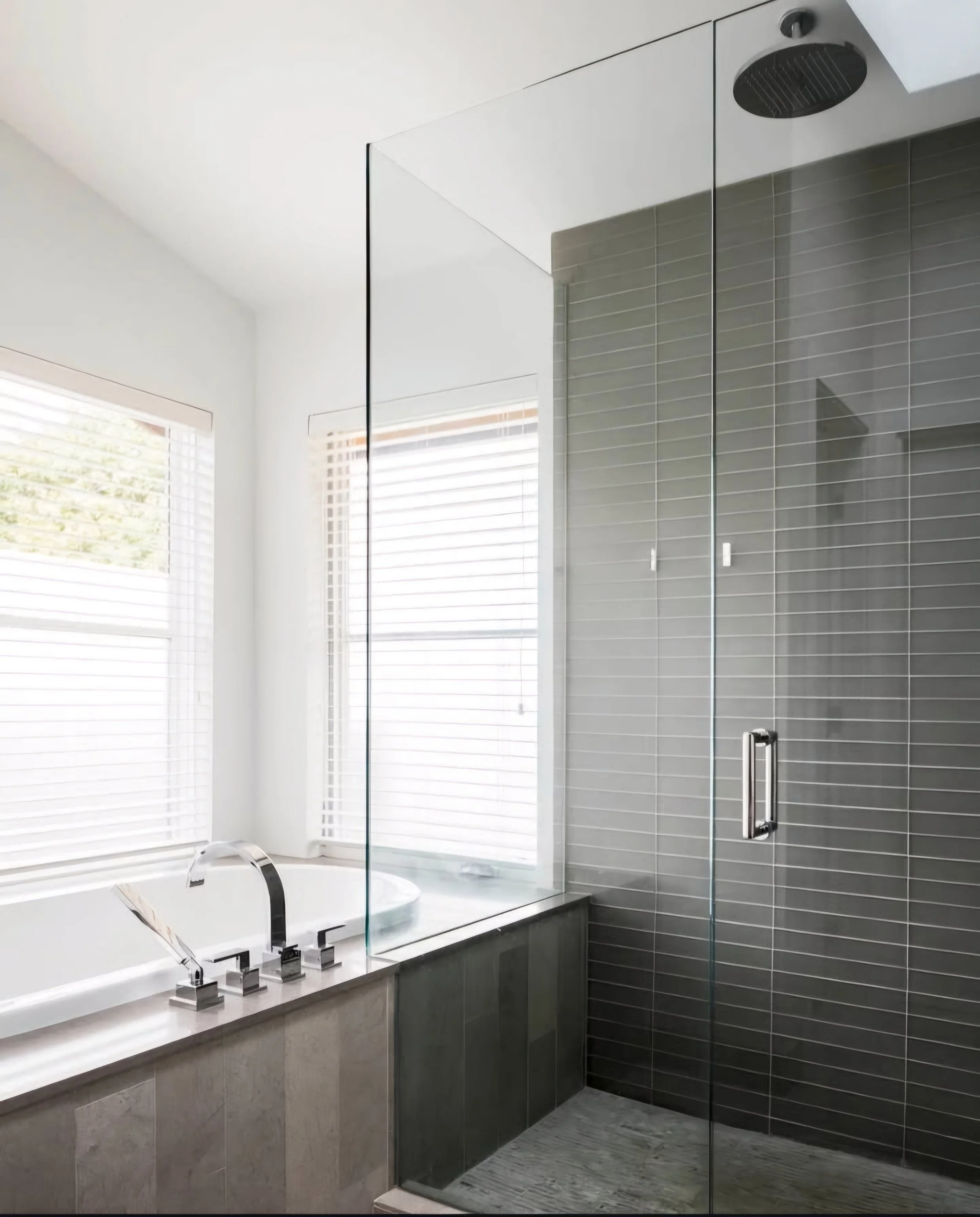Modern bathroom with a bathtub next to two windows with white blinds, and a glass shower enclosure with grey tile walls.