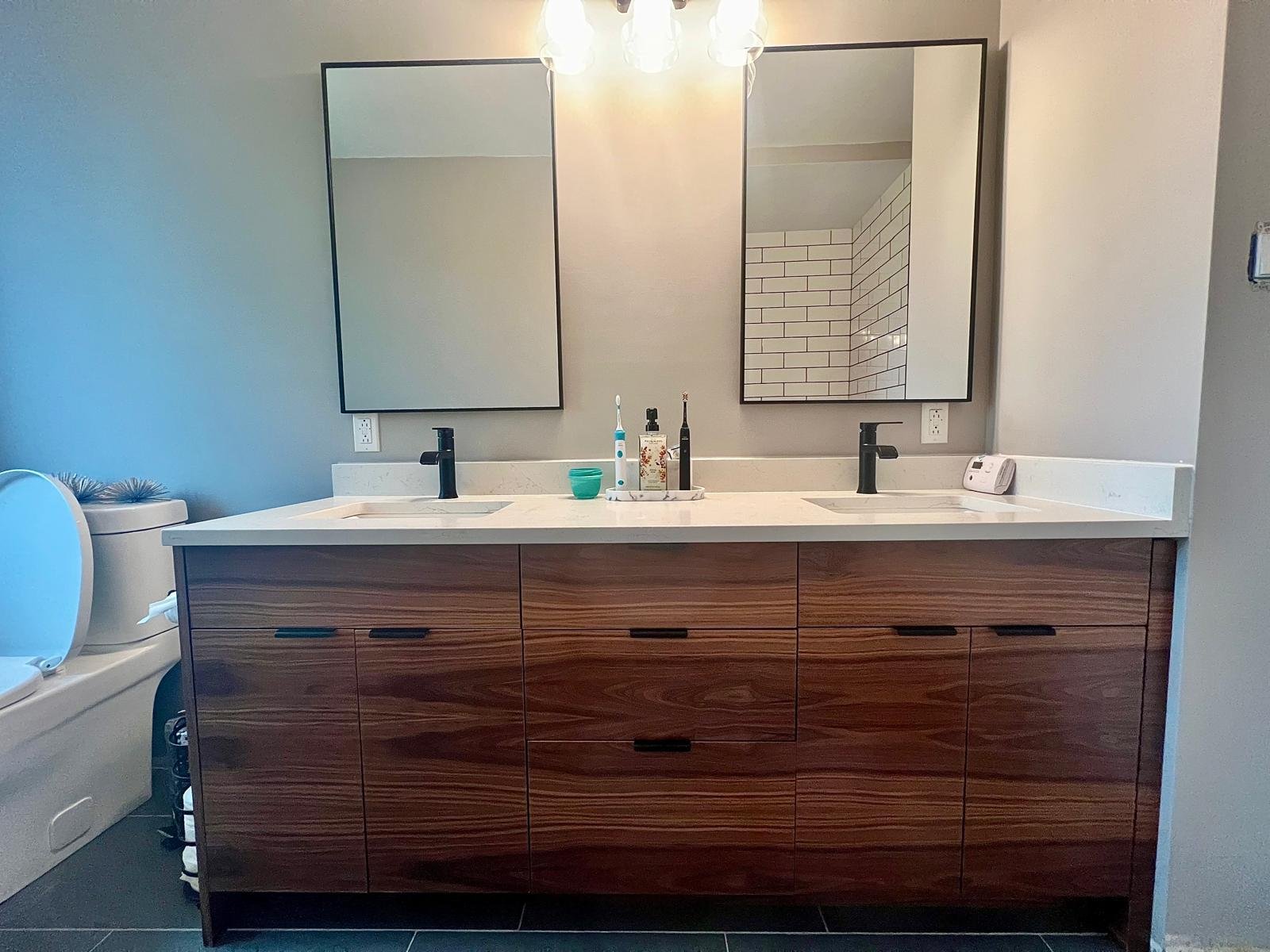 Modern bathroom with double vanity, wooden cabinet, two mirrors, toothbrush and soap on the counter, and a toilet to the left.