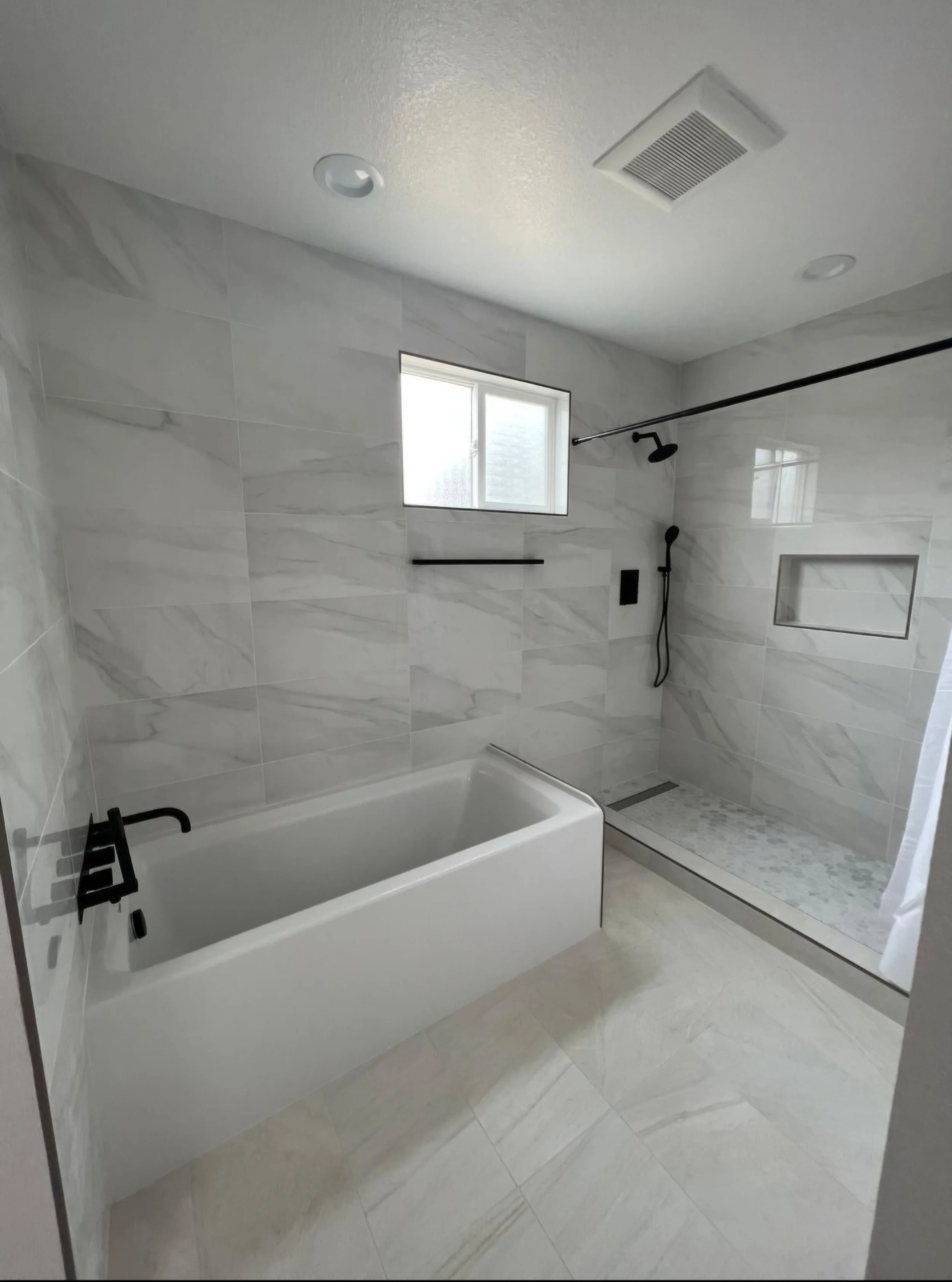Modern bathroom with bathtub and walk-in shower with black fixtures, gray marble tiles, small window, and recessed lighting.
