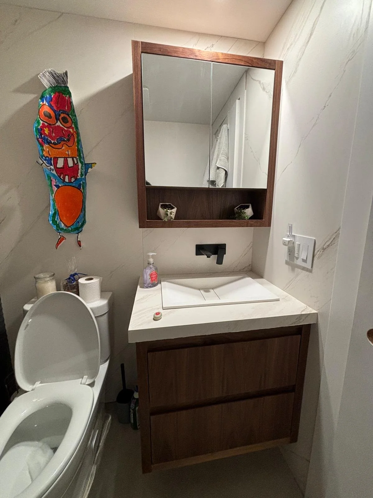 Small bathroom with a white toilet, a vanity with a white countertop, a rectangular sink, a black wall-mounted faucet, a wooden mirror cabinet, a hand soap dispenser, a small decorative planter, and a colorful paper lantern hanging on the wall.