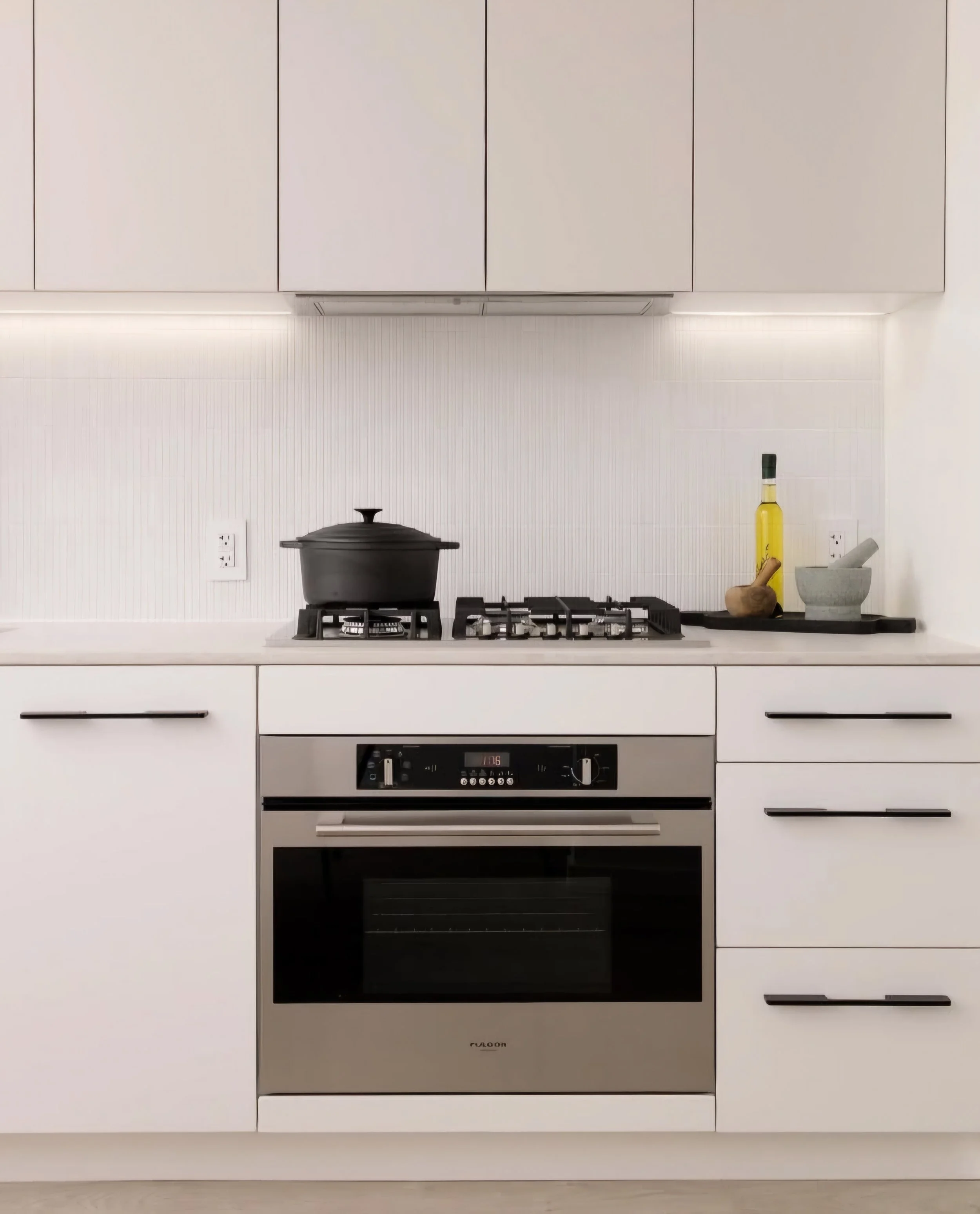 Minimalist white kitchen with a built-in oven, gas stove with a black pot, and countertop items including a yellow bottle, a garlic bulb, and a mortar and pestle on a black tray.