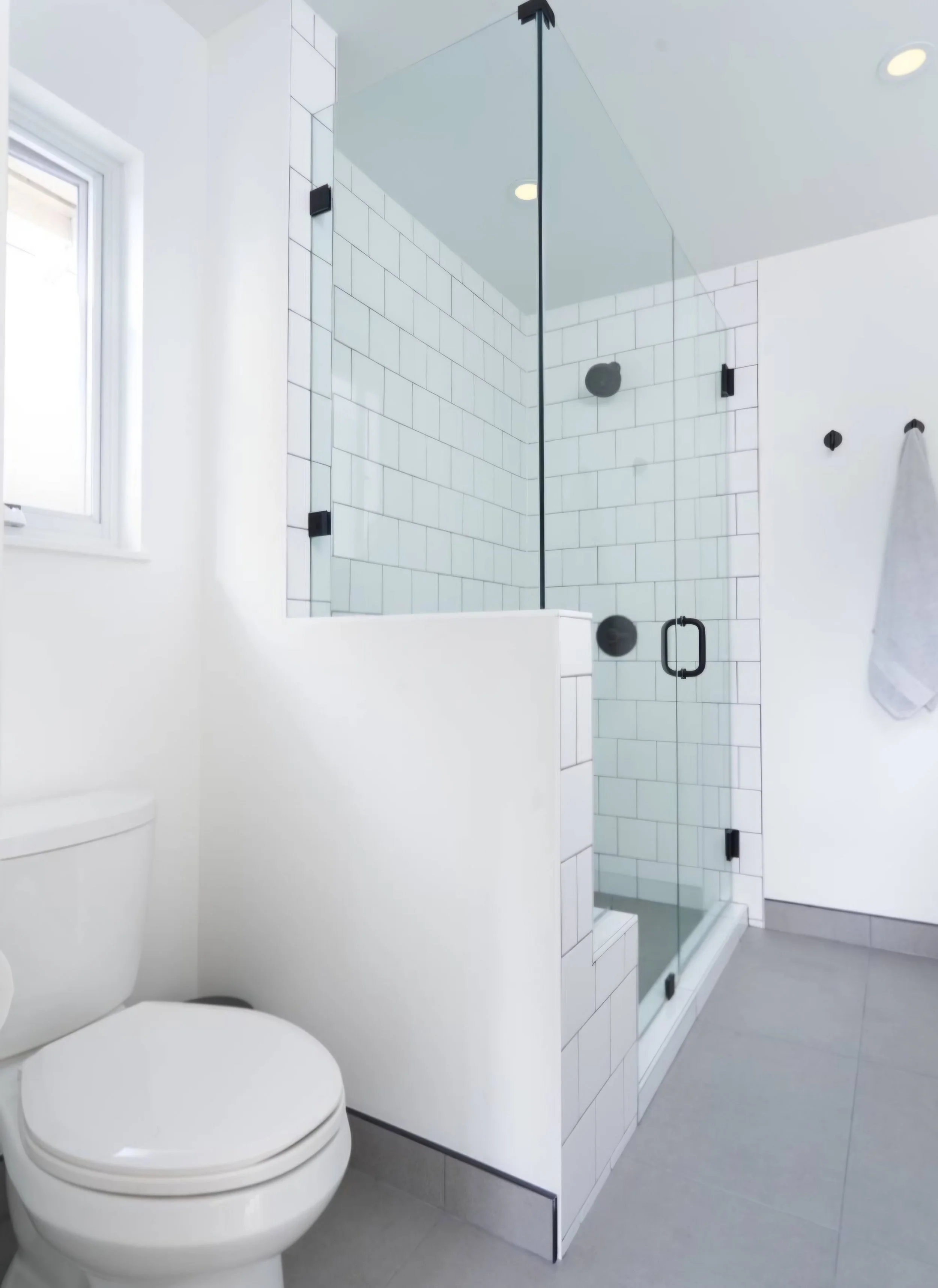 Modern bathroom with white walls, gray tiled floor, and a glass-enclosed shower with black fixtures. A white toilet is visible next to the shower, and a window allows natural light into the space. A towel hangs on a hook on the wall.