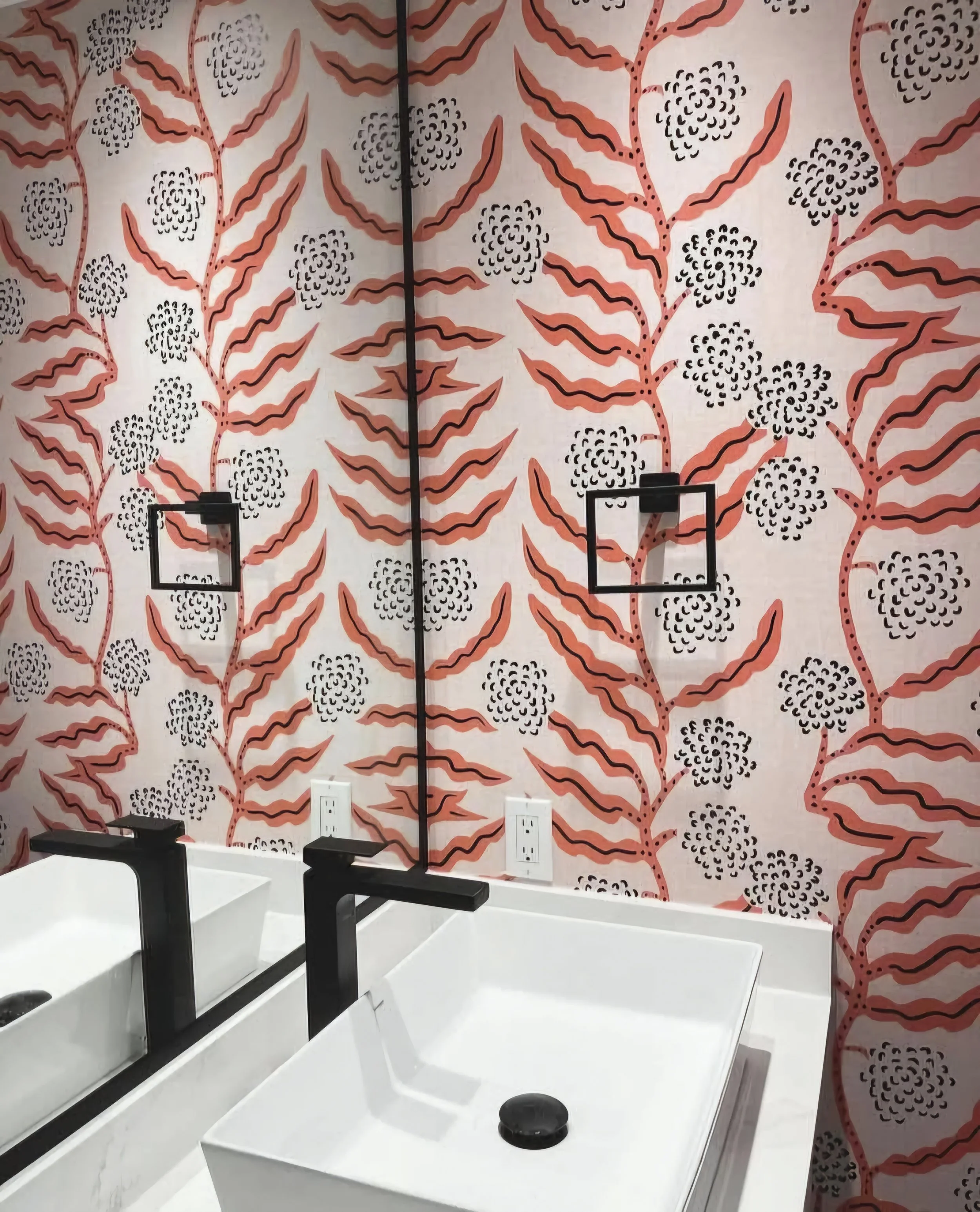 Bathroom sink with black faucet and white square basin, mirror with black frame, patterned wallpaper with red and black abstract floral design, and two electrical outlets.