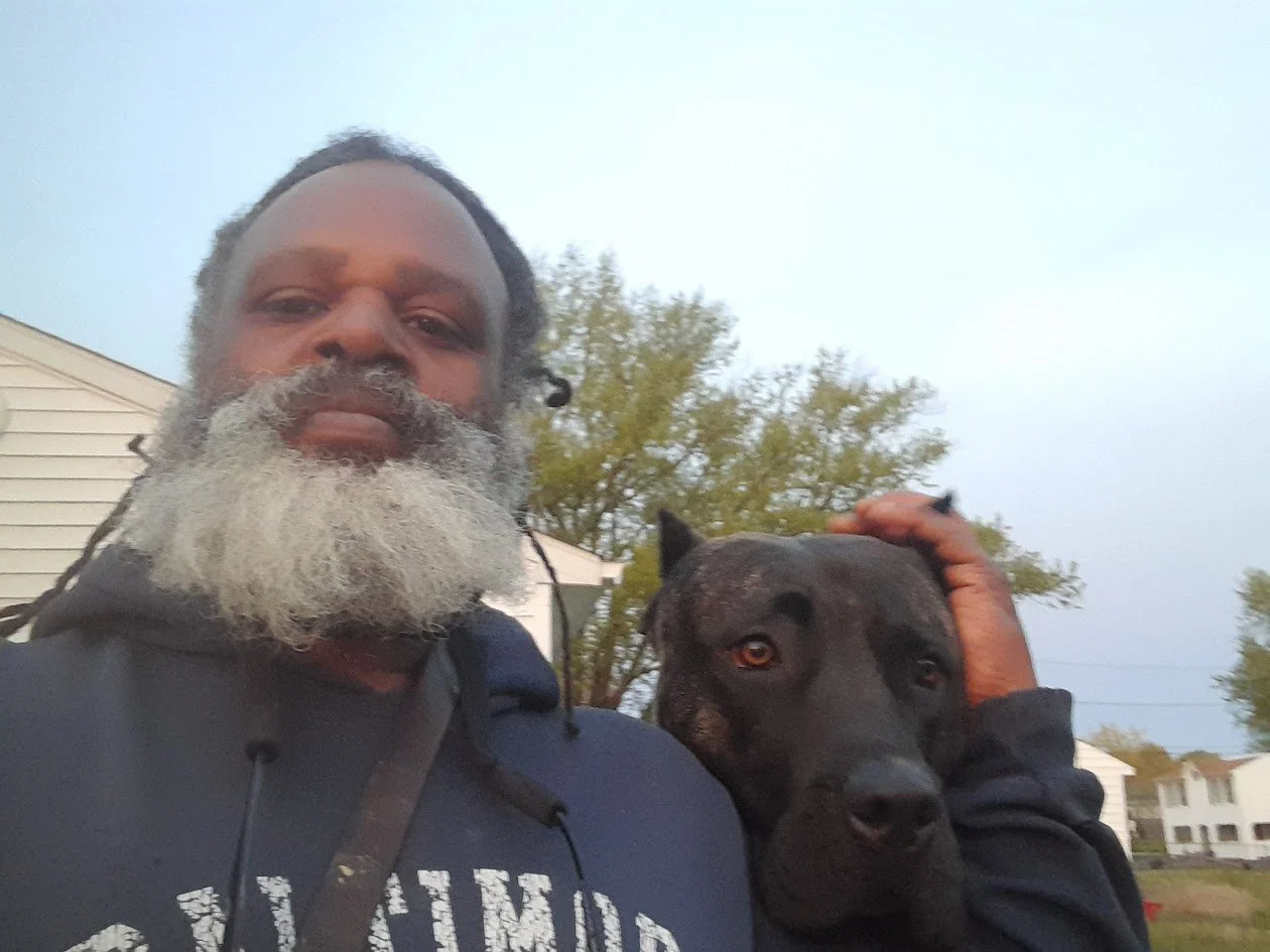 A man with a gray beard taking a selfie with a black dog outdoors during daytime.