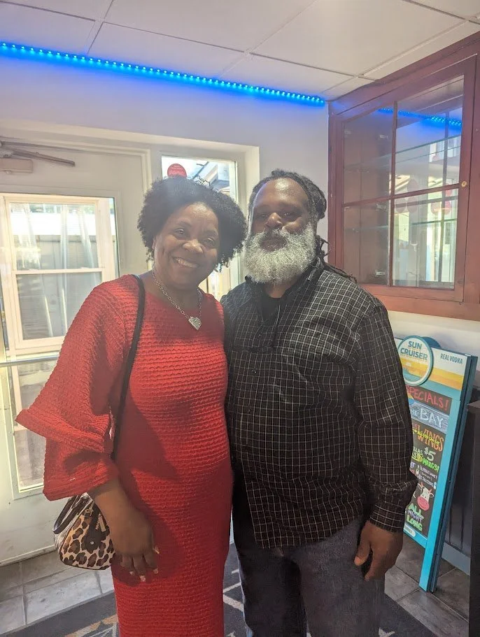 A smiling woman wearing a red dress and a man with gray beard in a black checkered shirt standing indoors near a door and window, with a promotional stand and blue LED lights overhead.