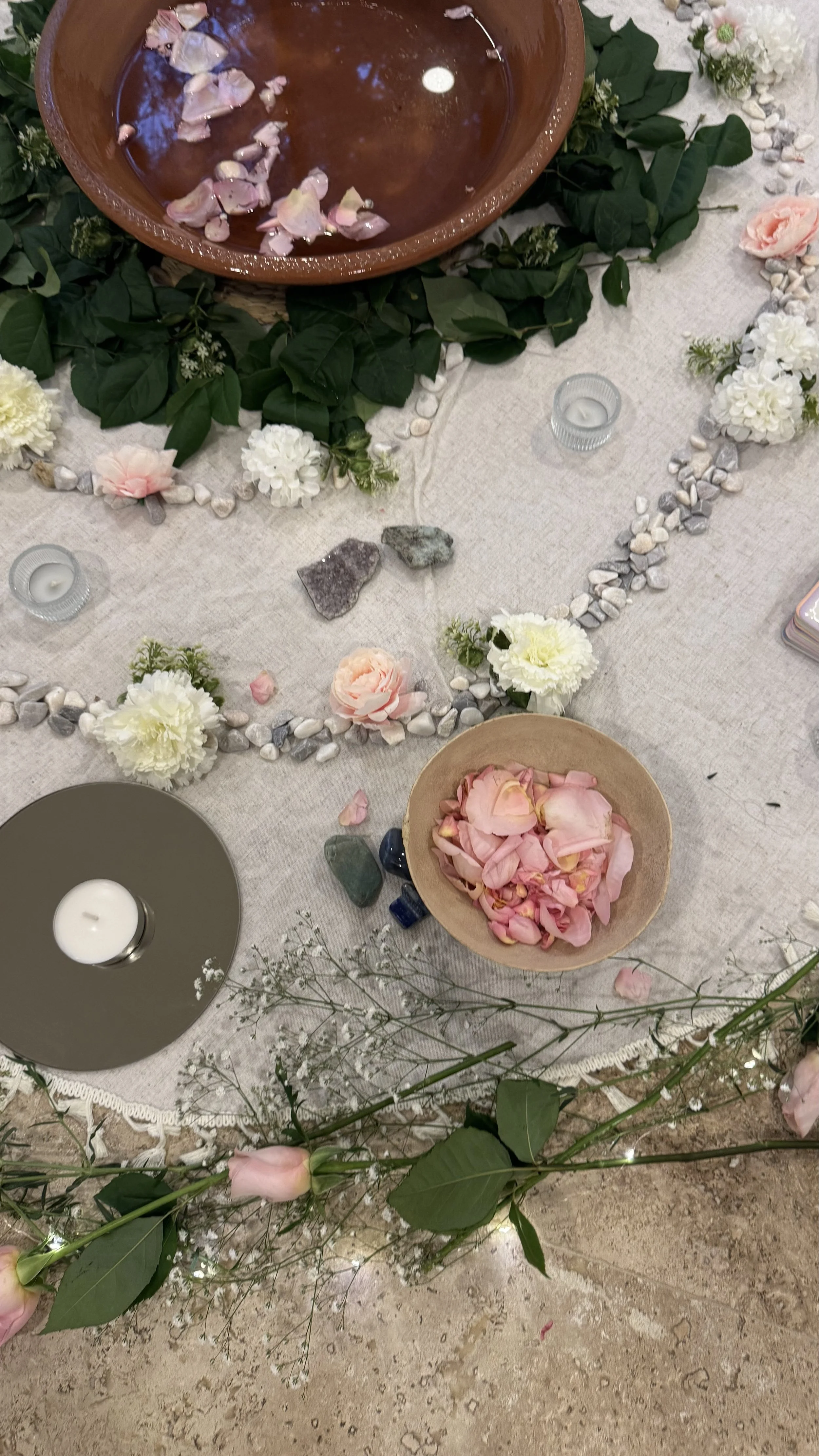 Mesa decorada con flores, piedras y velas, con un recipiente de cerámica grande con agua y pétalos, y un tazón pequeño con pétalos de rosa.