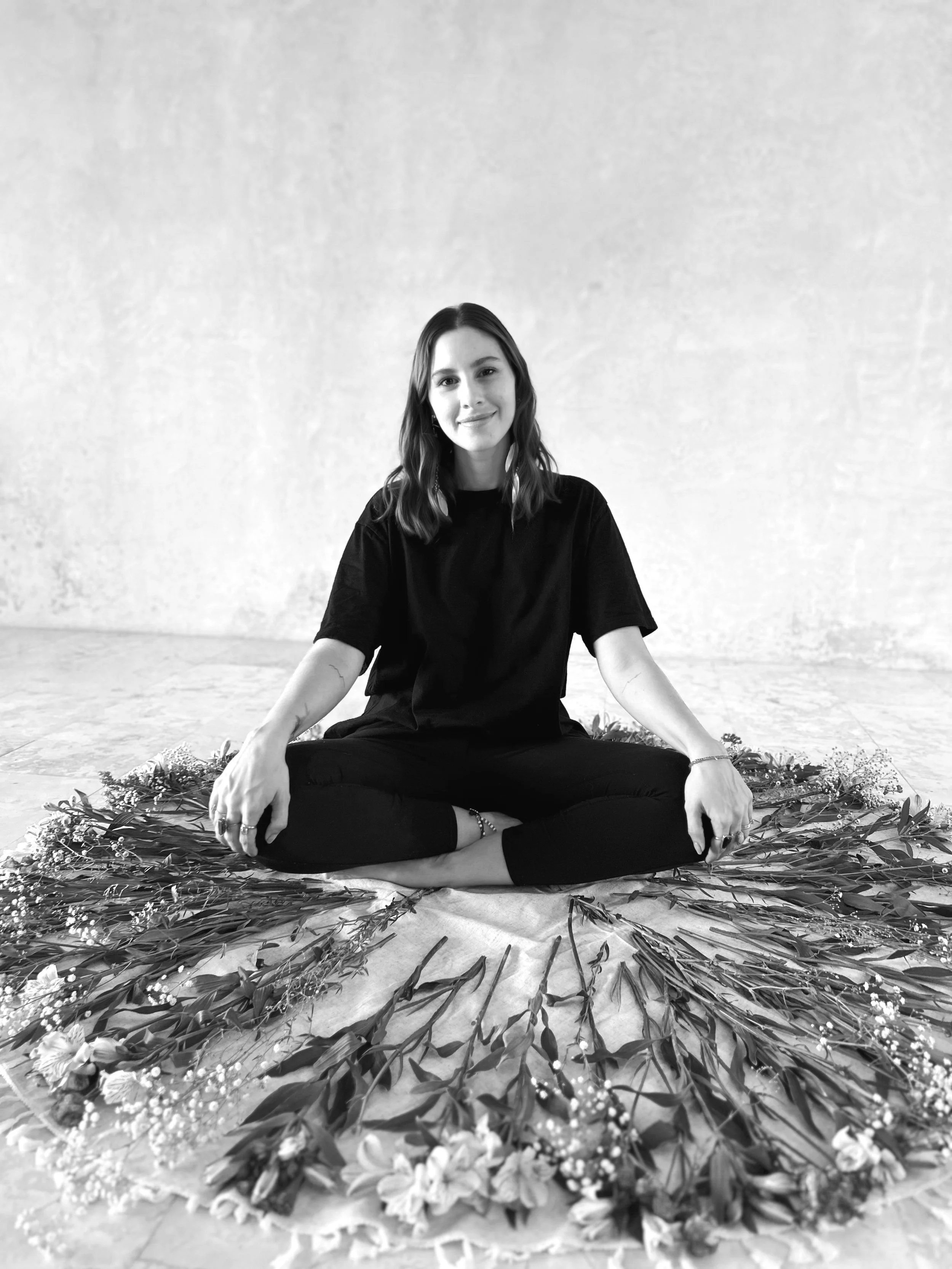 Retrato en blanco y negro de una joven mujer chamana, holística, en una ceremonia holística, con una expresión serena, rodeada por arreglos florales en un círculo en el suelo, en un ambiente holístico y luminoso.