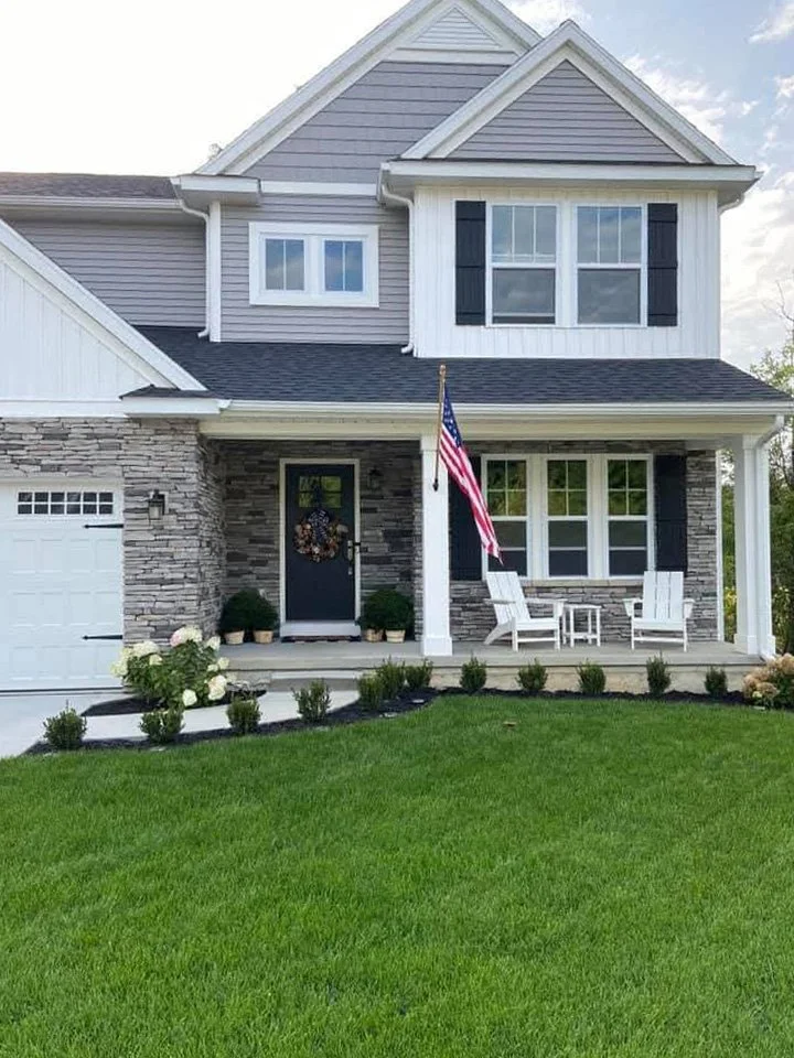 A two-story house with gray siding, white trim, and black shutters, featuring a front porch with white chairs, an American flag, and a well-manicured, lush lawn in a residential suburb in Michigan.