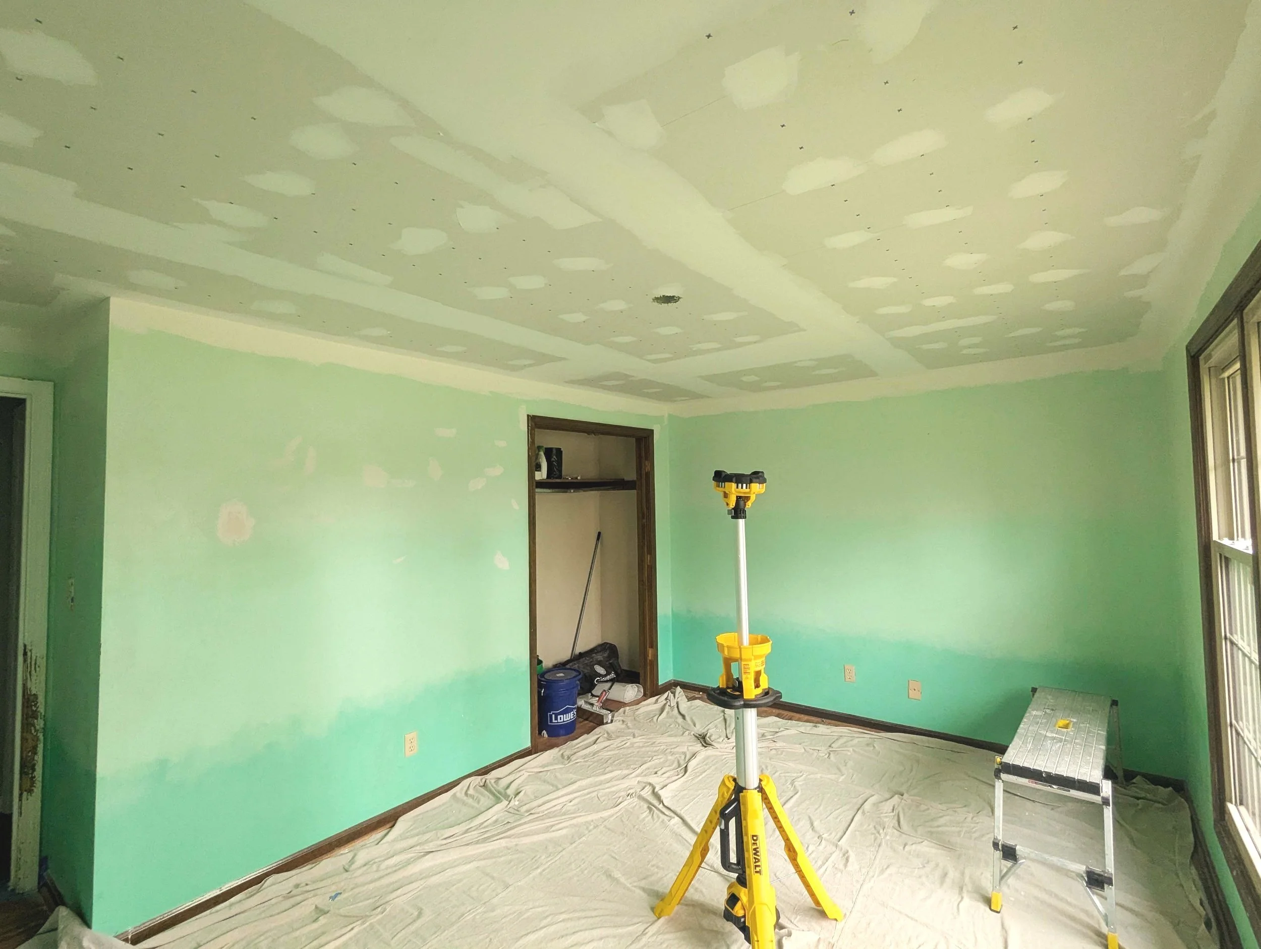 Room under renovation with green painted walls, unfinished ceiling with drywall patches, and construction tools including a yellow support stand and a step ladder on a plastic-covered floor.