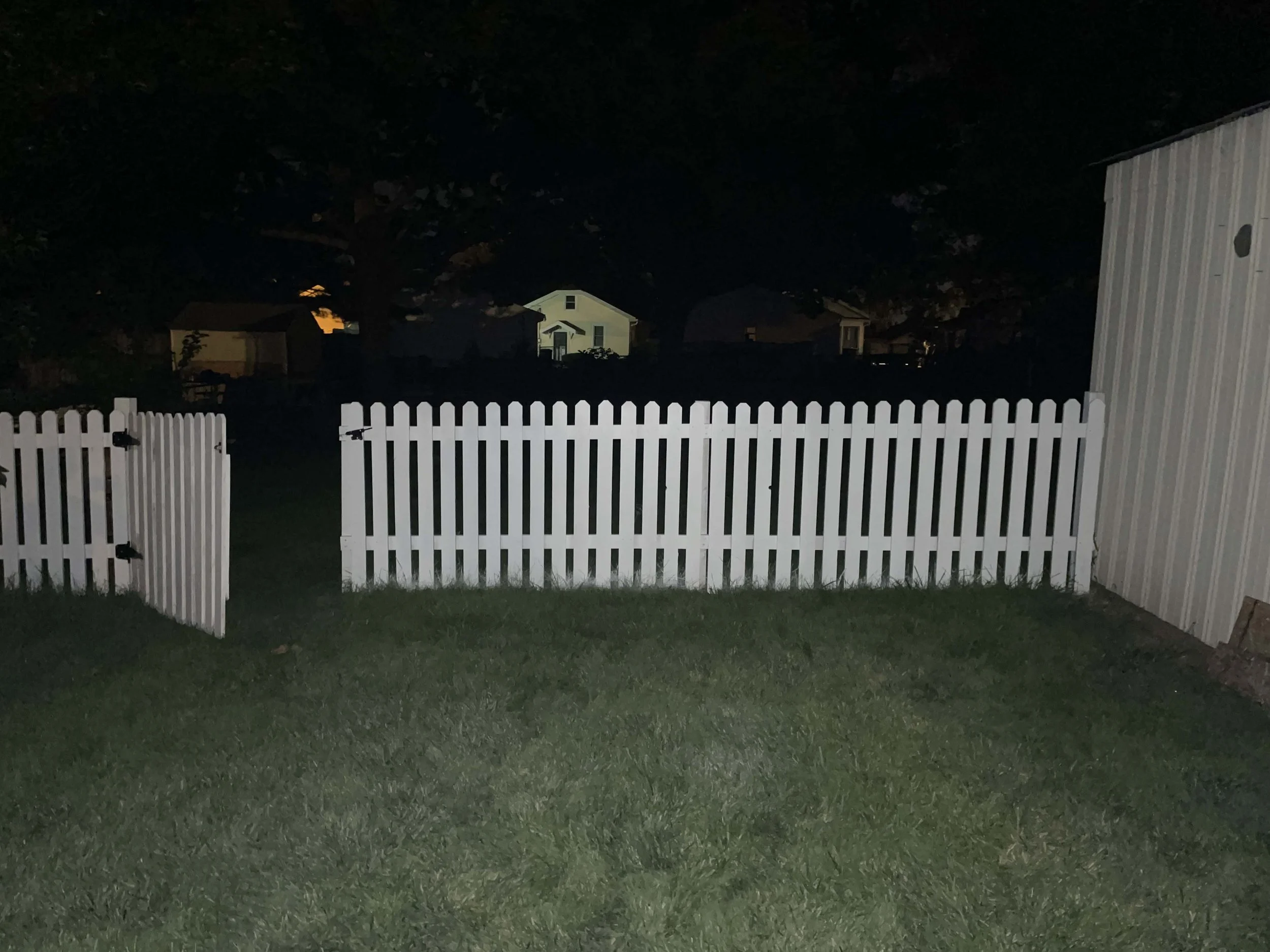 Nighttime view of a backyard with a white picket fence, an open gate, a grassy lawn, and a small shed on the right side. Houses are visible in the distance with trees and darkness overhead.