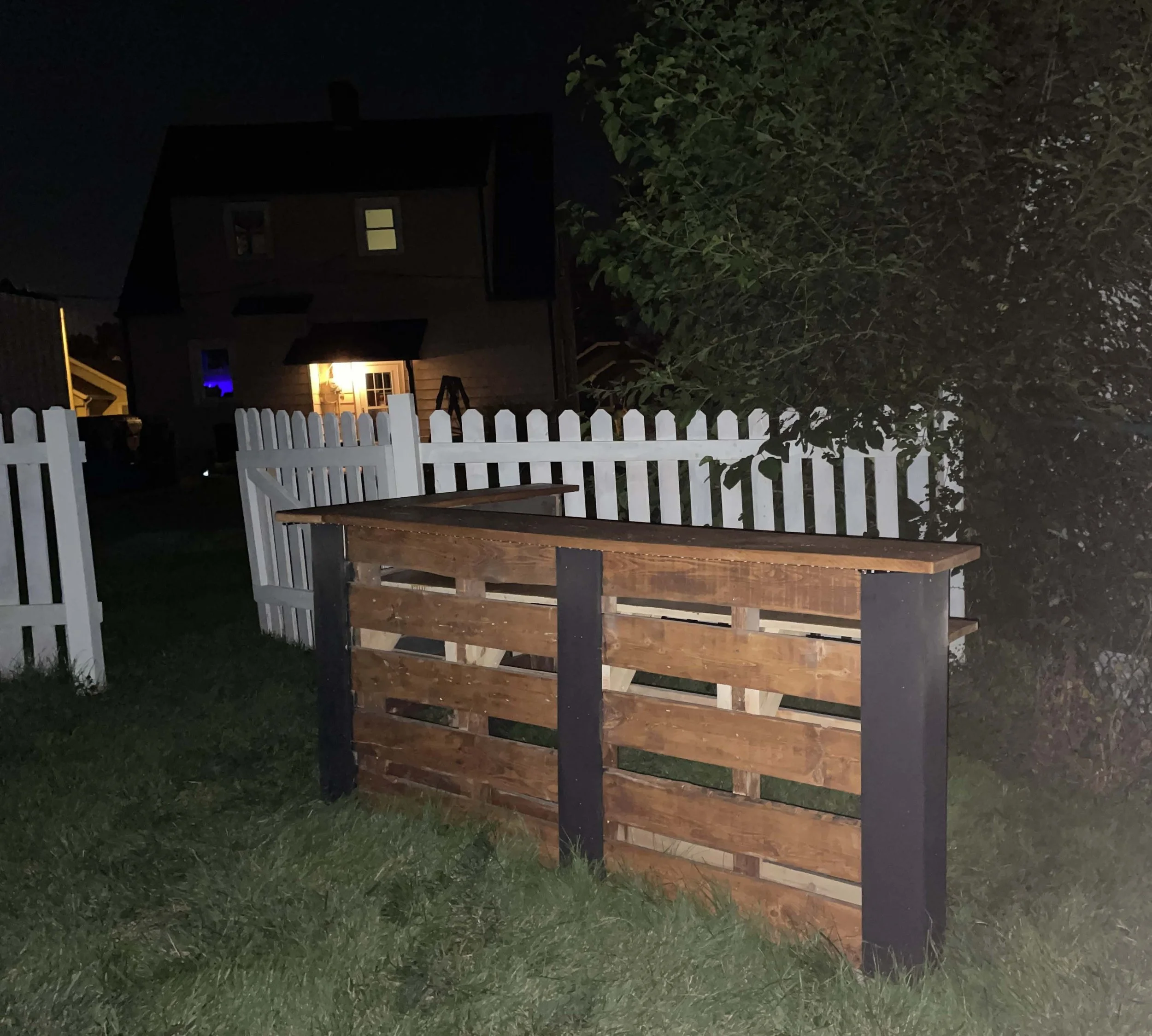 Nighttime photo of a wooden planter box with a dark house and lit windows in the background, white picket fence and green grass in the yard.