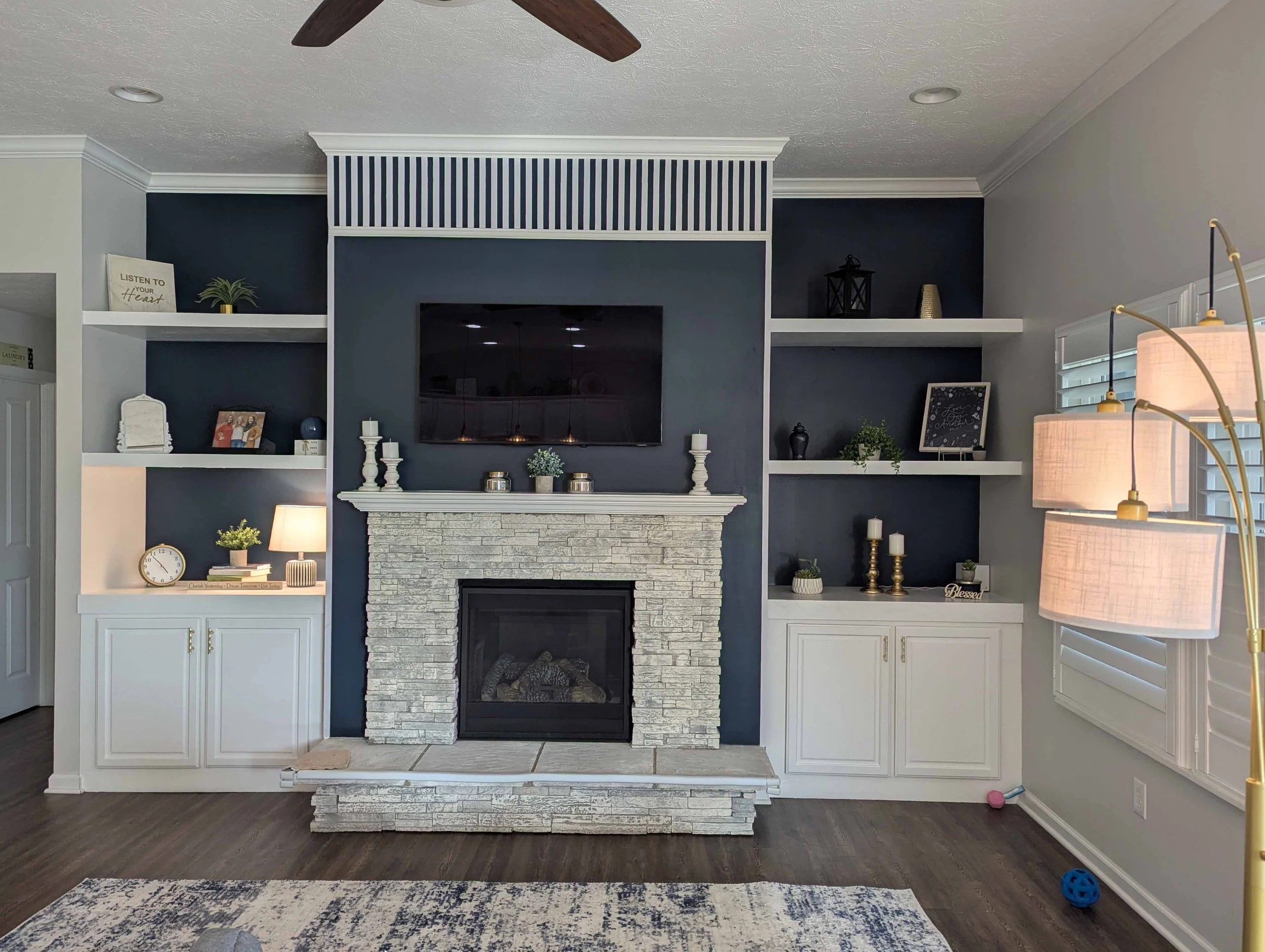 Living room with a stone fireplace, built-in shelves on each side, a flat-screen TV above the fireplace, white cabinetry, decorative items, a window with white shutters, a blue and white area rug, and a multi-shade floor lamp.