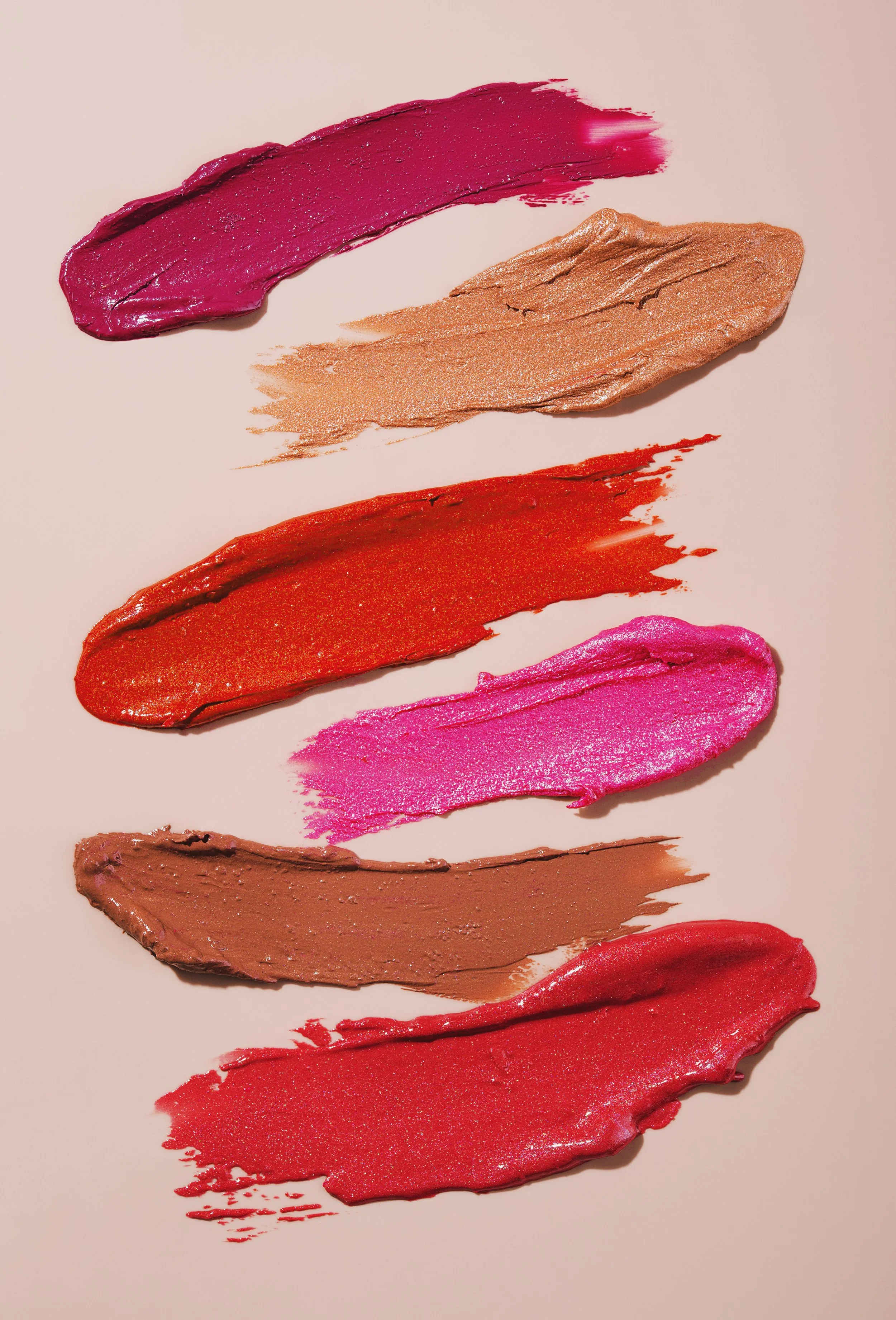 Swatches of various shades of lipstick or cosmetic cream on a light background, including purple, beige, red, pink, brown, and orange.
