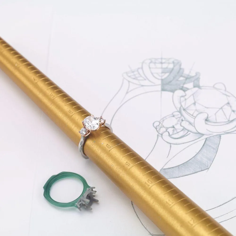 A diamond ring placed on a gold measurement tool, with a jewelry design sketch and a small green repair tool nearby.