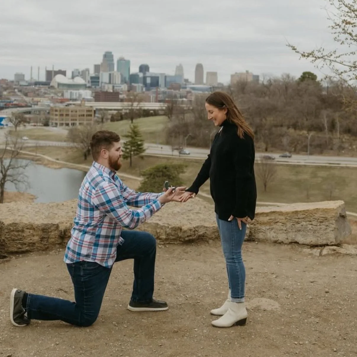 A man on one knee proposing to a woman outdoors with city skyline in the background.
