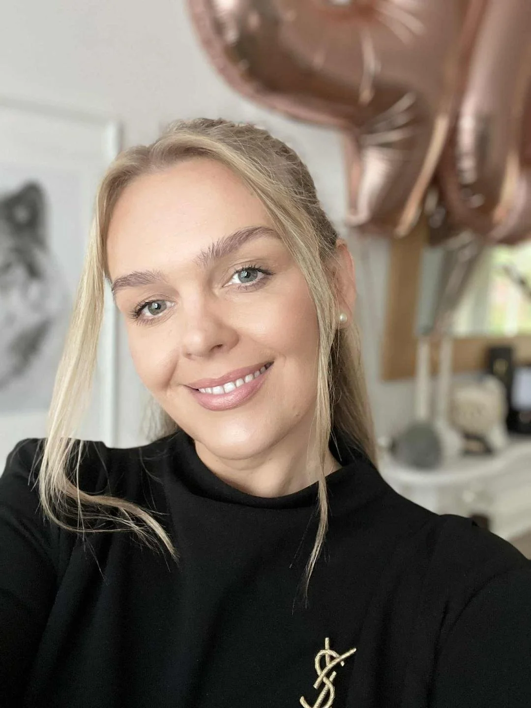 A woman taking a selfie indoors, smiling with blonde hair, wearing a black top, pearl earrings, and a gold jewelry pendant. In the background, there are rose gold balloons, a blurred wolf picture, and a bookshelf.