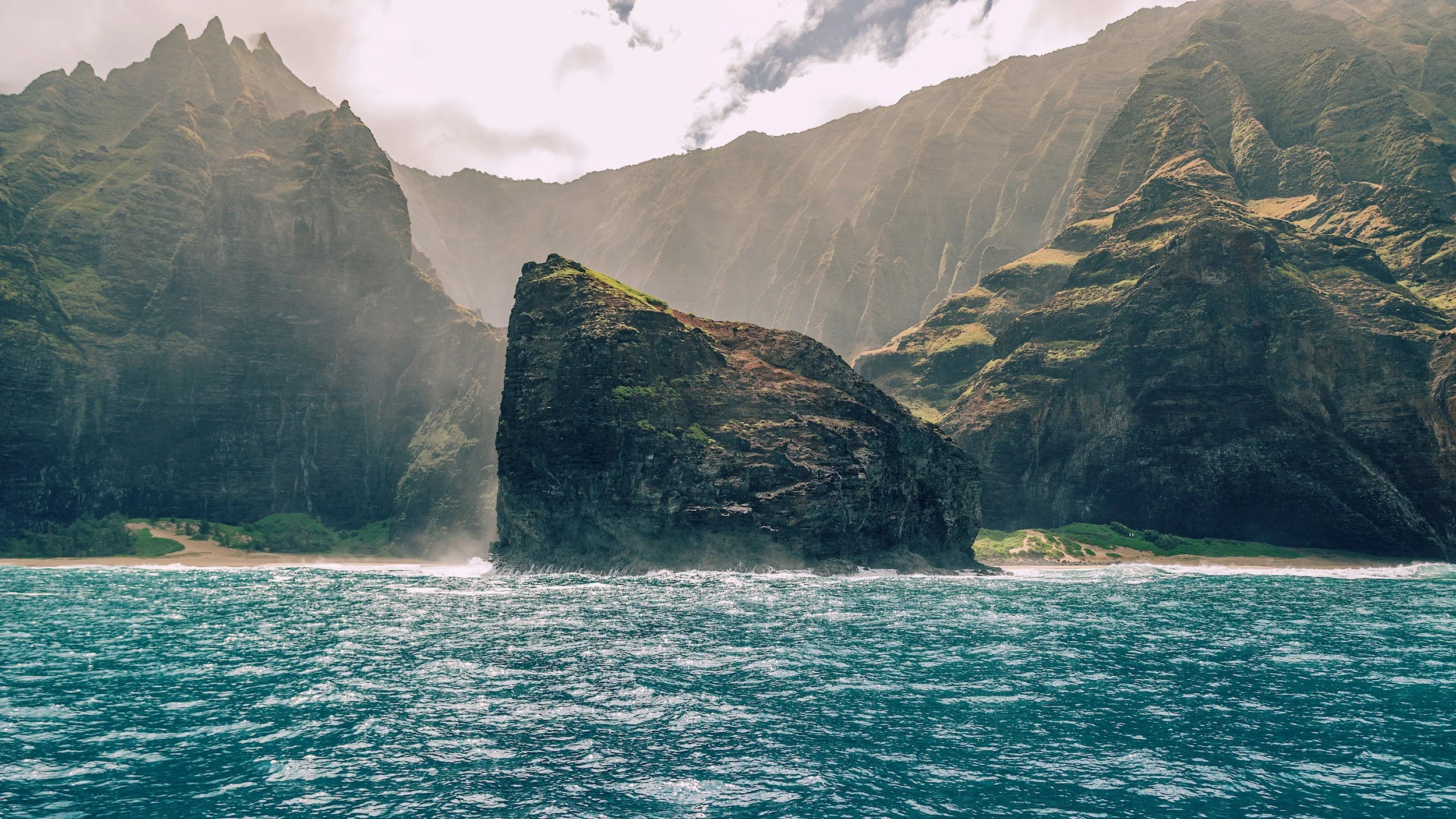 A large, dark rock formation rising from the ocean near a mountain cliff. The water is turquoise with waves, and the mountains are partly covered in greenery, with mist and clouds overhead.