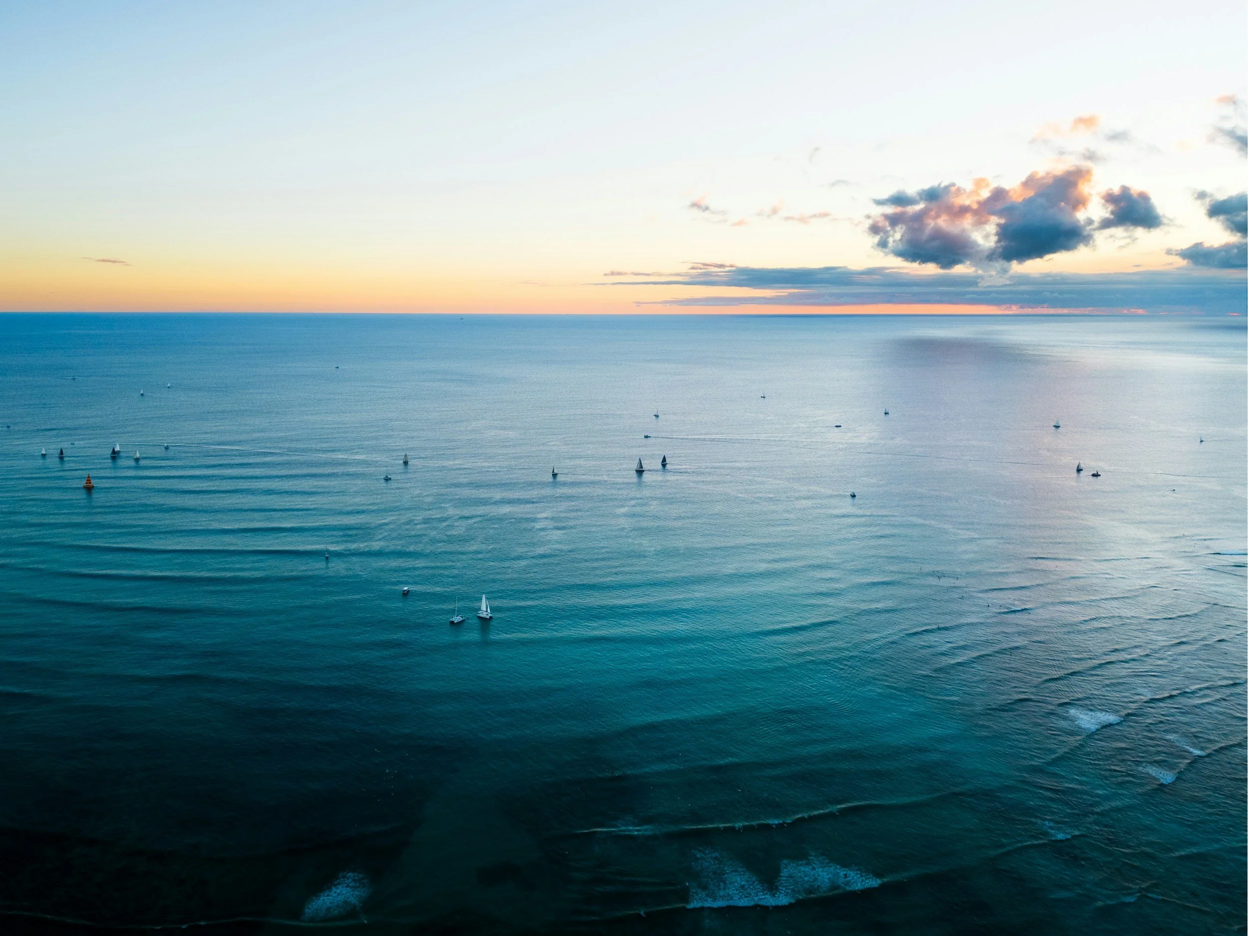 Ocean view at sunset with sailboats and clouds in the sky.
